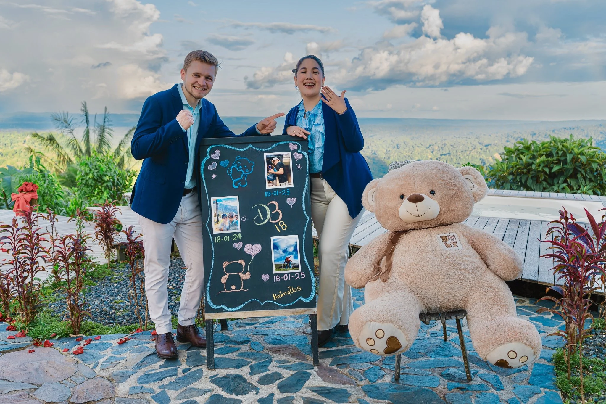 Una pareja con ropa formal azul y blanca posando frente a una pizarra con fotos y fechas, rodeados de plantas, con un oso de peluche gigante y un fondo de paisaje montañoso y cielo con nubes.