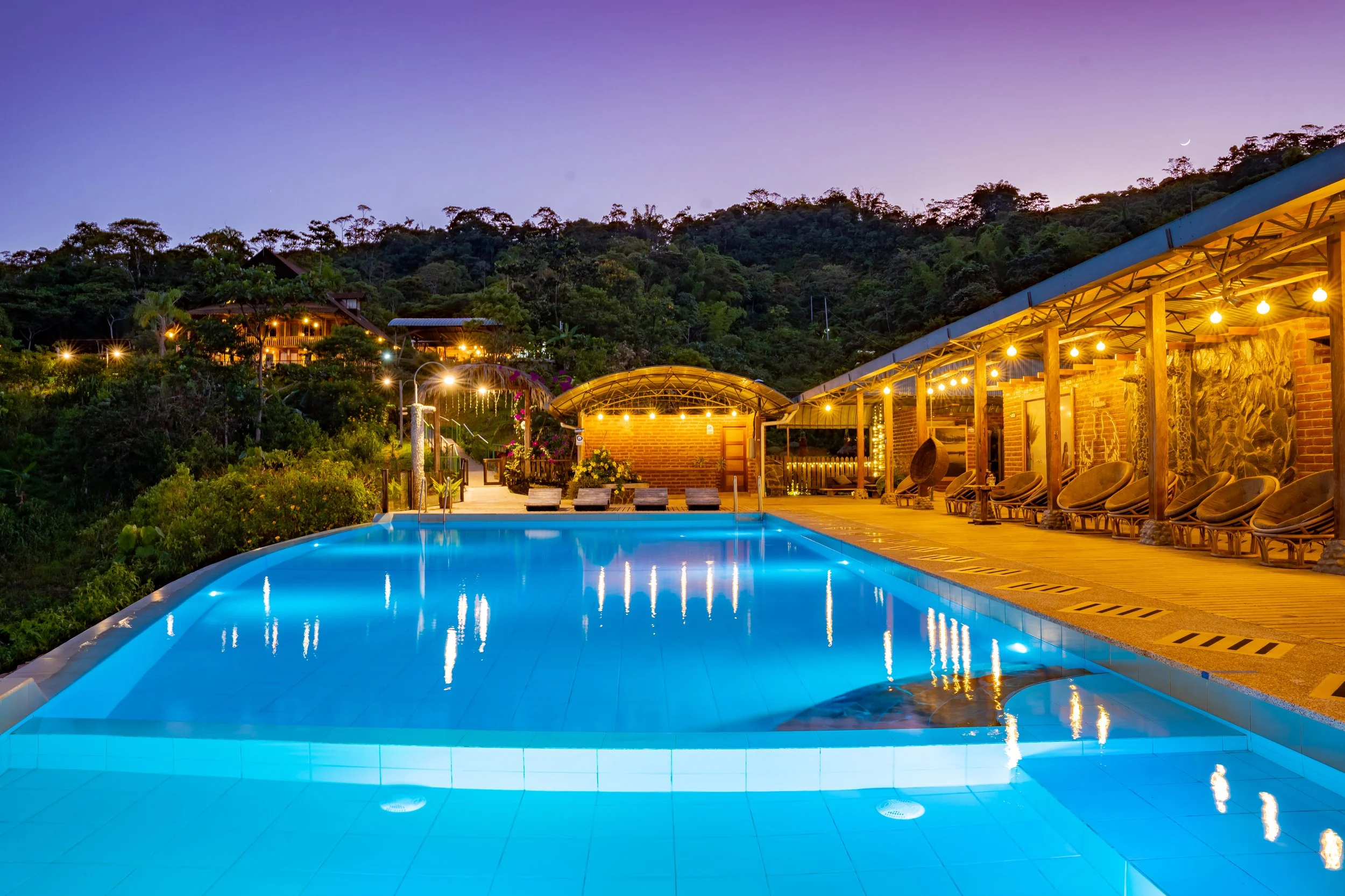 Piscina iluminada con luces cálidas, rodeada de una terraza con sillones y una estructura de madera, en un entorno montañoso con casas en la colina, al atardecer.