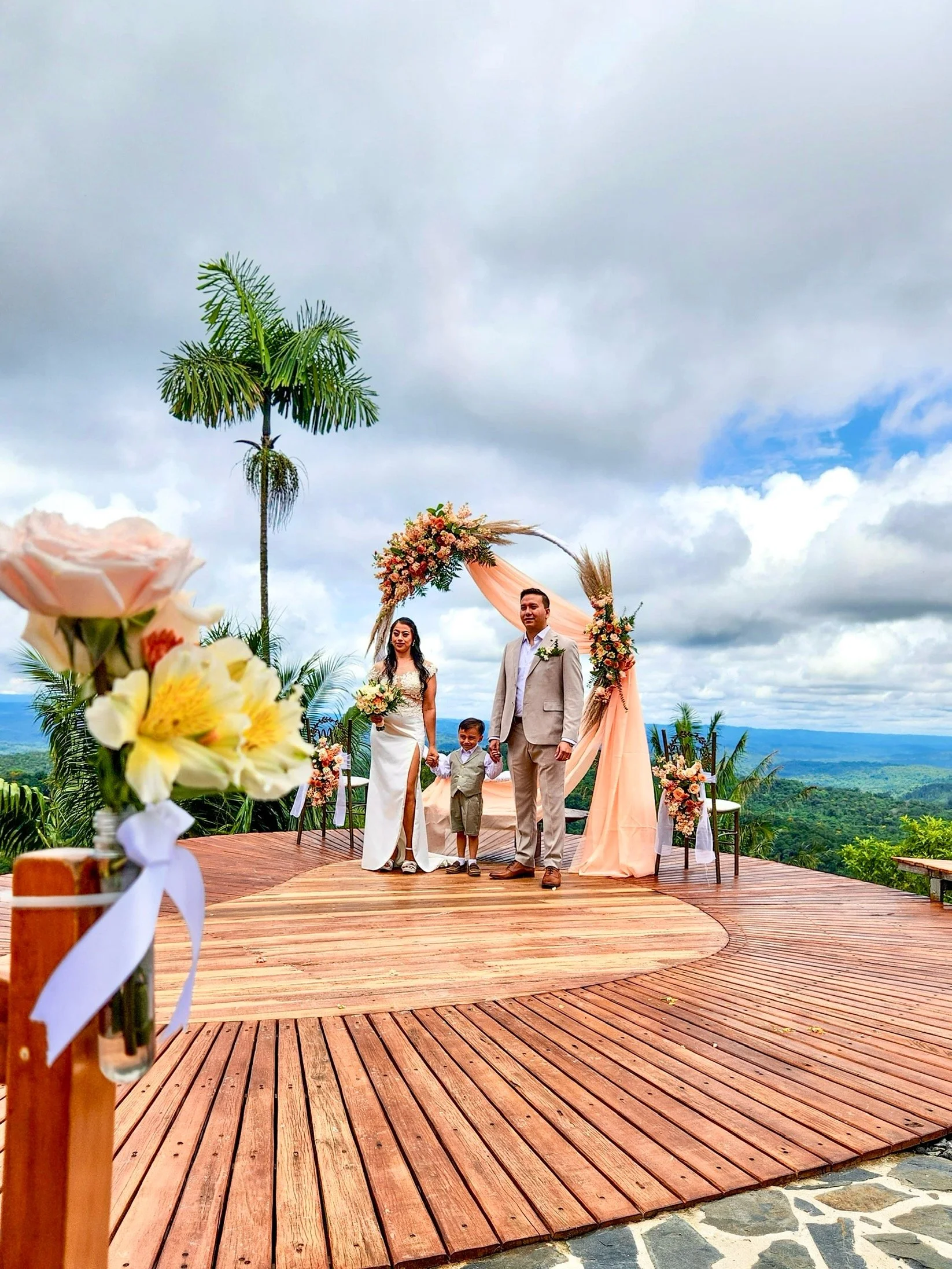Pareja de novios con un niño en una ceremonia de boda al aire libre, decorada con flores y rodeada de naturaleza y cielo nublado.