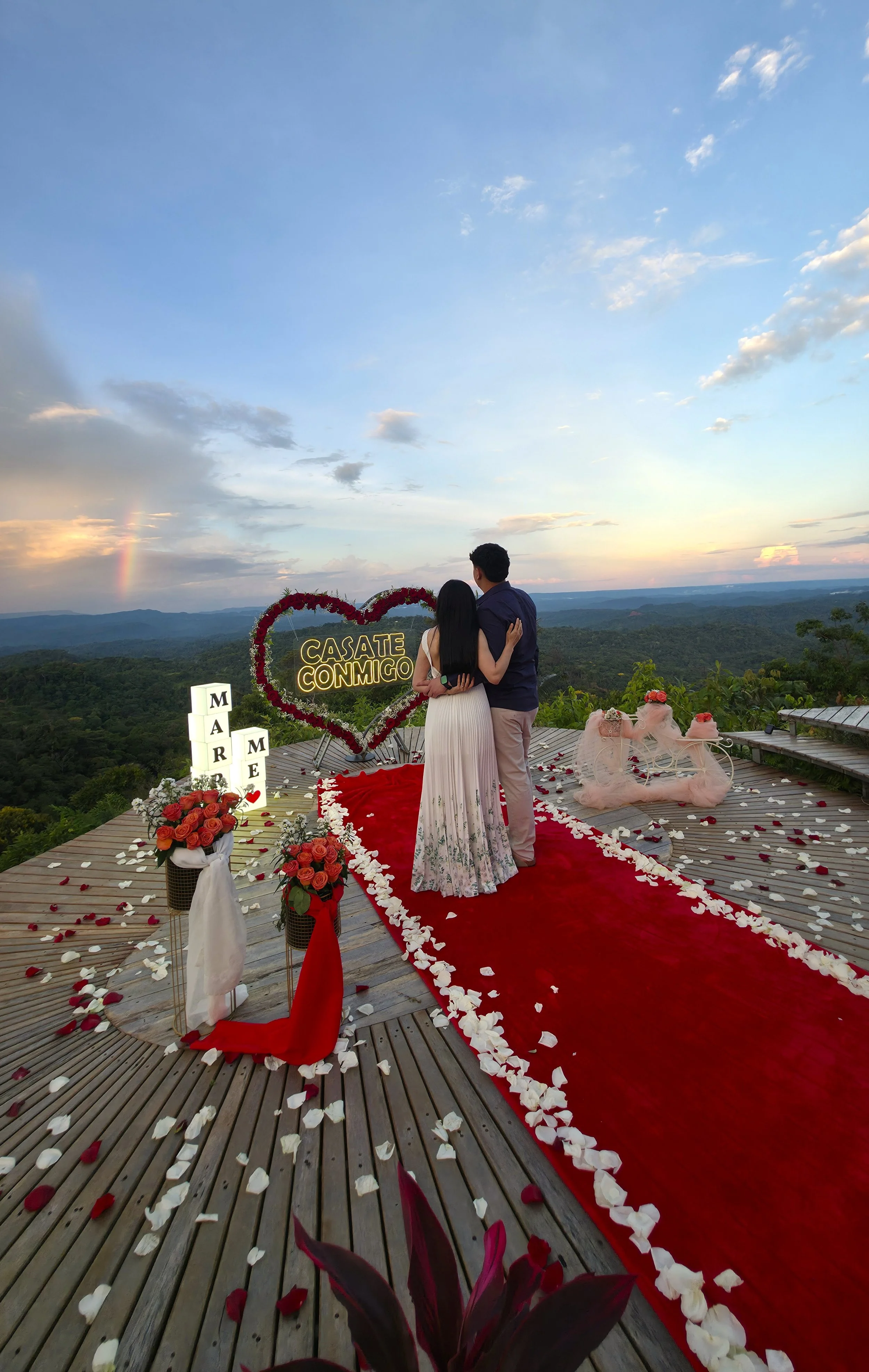 Pareja en una ceremonia romántica en un mirador con vista a la naturaleza, decorada con flores y corazones, en el atardecer.