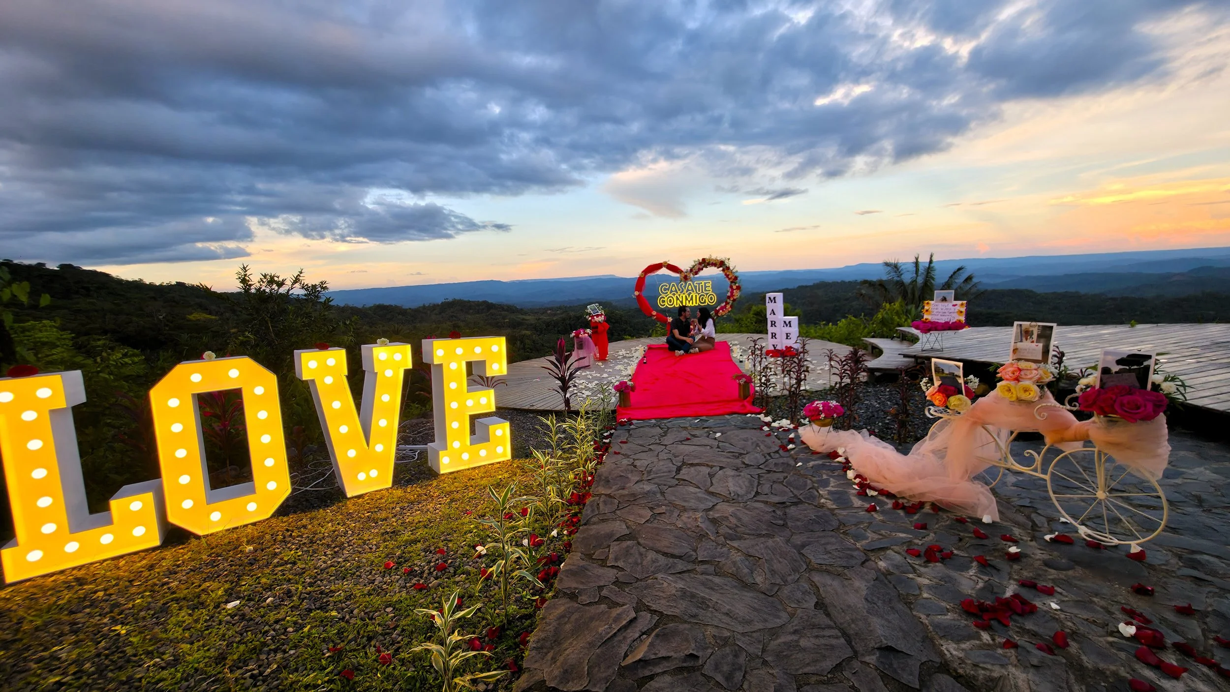 Decoración de pedidad de mano al aire libre con letras luminosas que dicen 'LOVE', un corazón con la inscripción 'CASATE CONMIGO', una pareja en el altar, un carruaje decorado con flores y fotos, y sendero cubierto de pétalos de rosa.