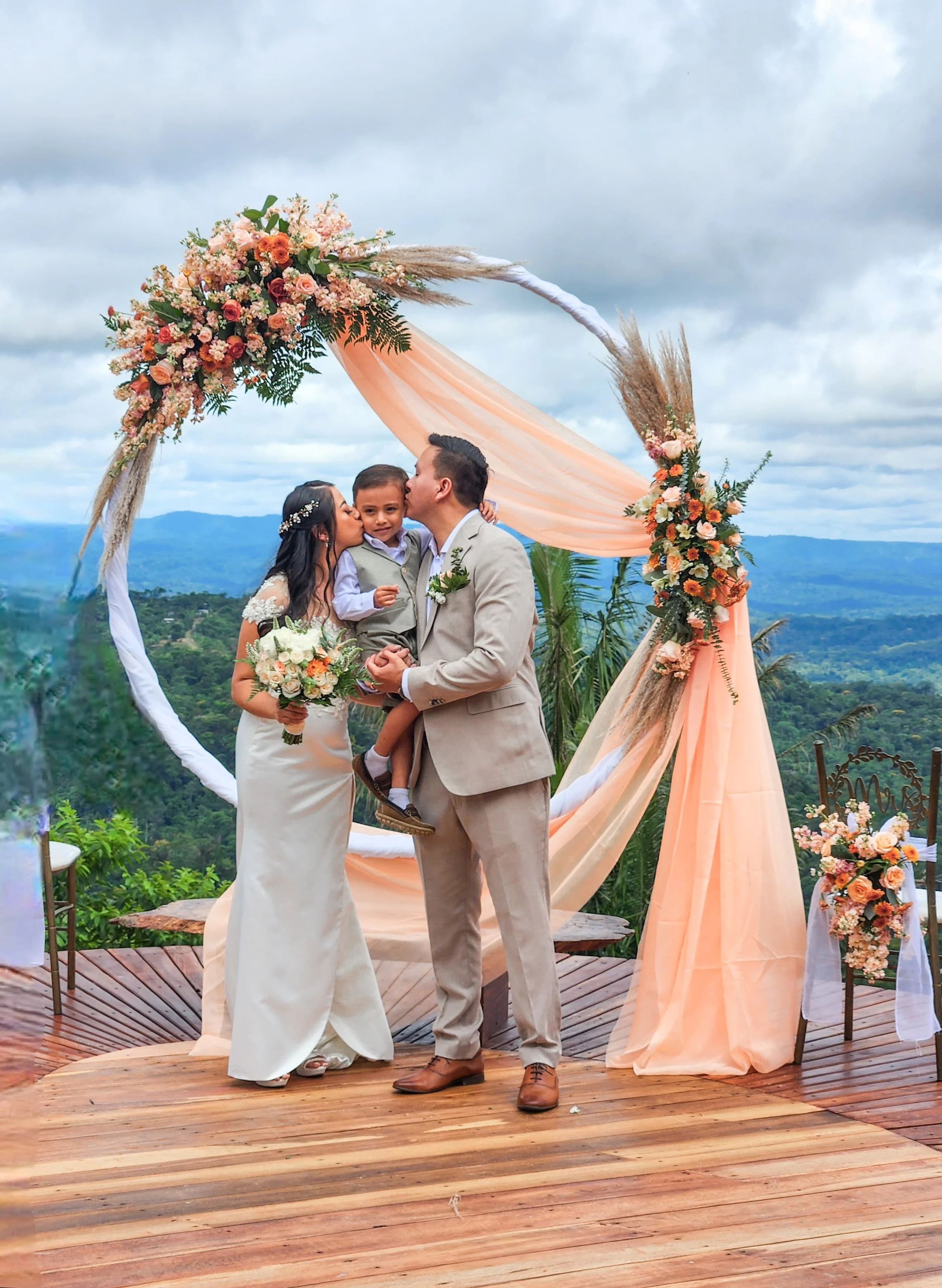 Pareja de novios y un niño en ceremonia de bodas al aire libre con decoraciones florales y un fondo de montañas y cielo nublado.