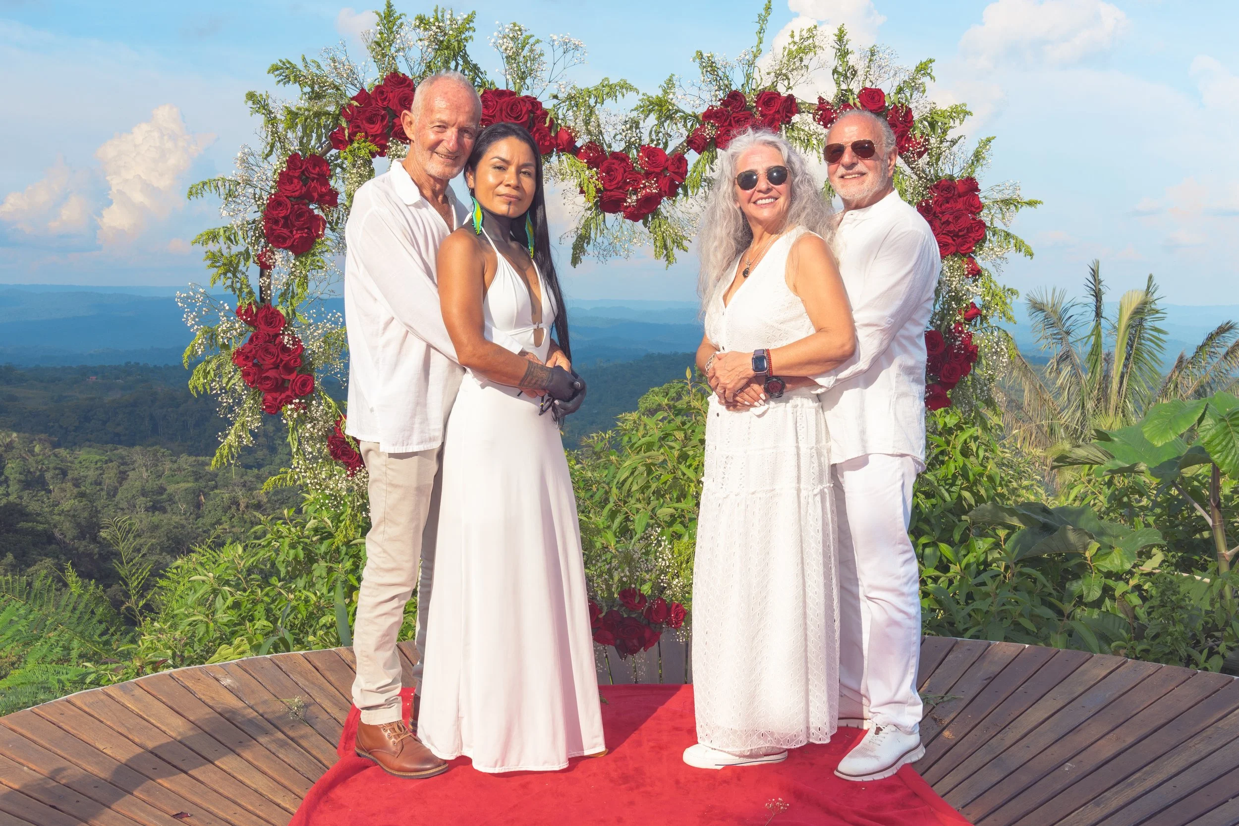 Grupo de cuatrp personas en boda al aire libre con fondo de montañas y cielo azul, rodeados de flores y decoraciones rojas, todos vestidos en colores blancos.