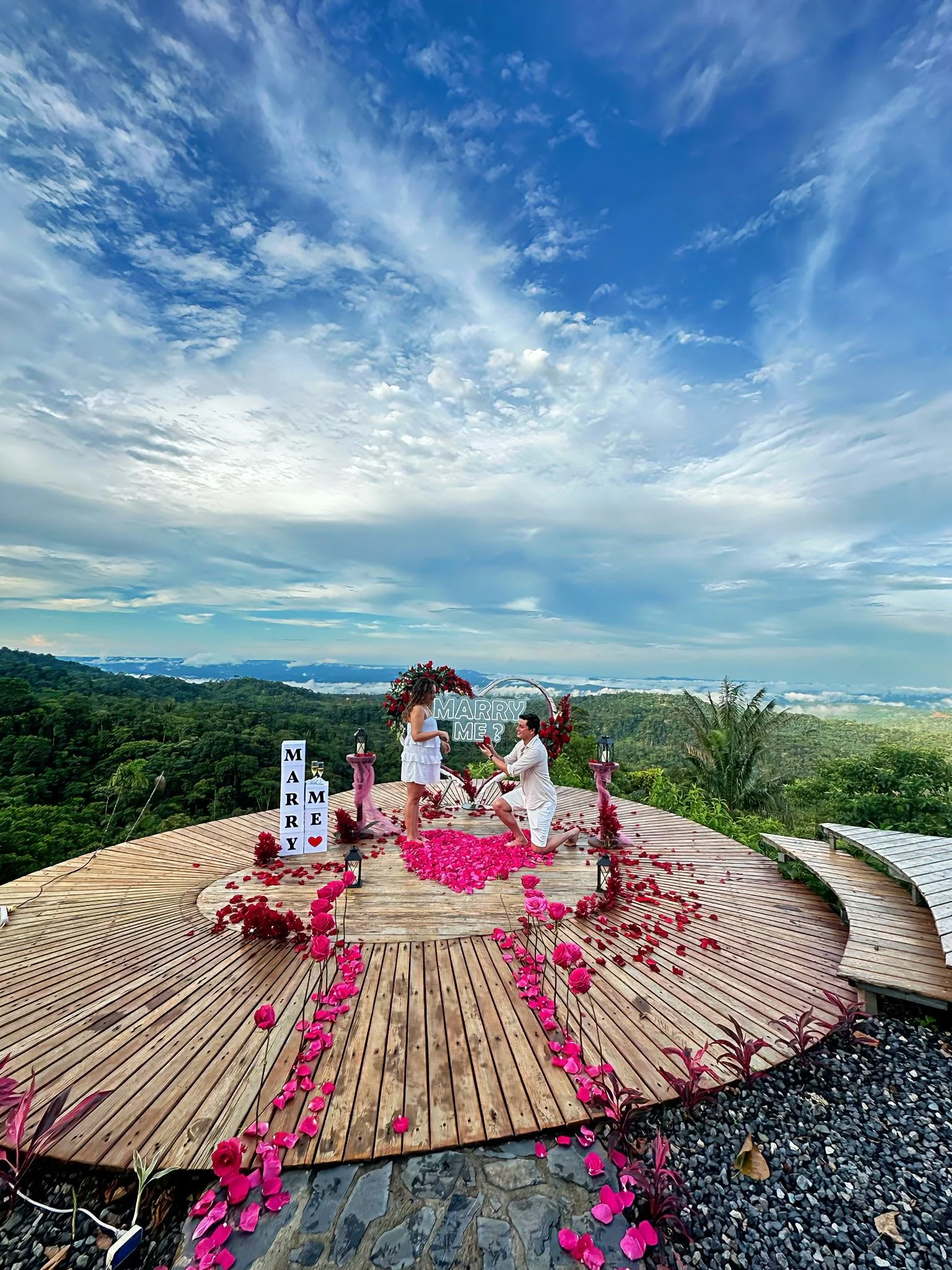 Una propuesta de boda en una plataforma de madera elevada en medio de un paisaje montañoso, decorada con muchas flores rosas y rojas, con una pareja en medio, uno arrodillado y la mujer con un anillo, y un cartel que dice "Marry Me" en el fondo.