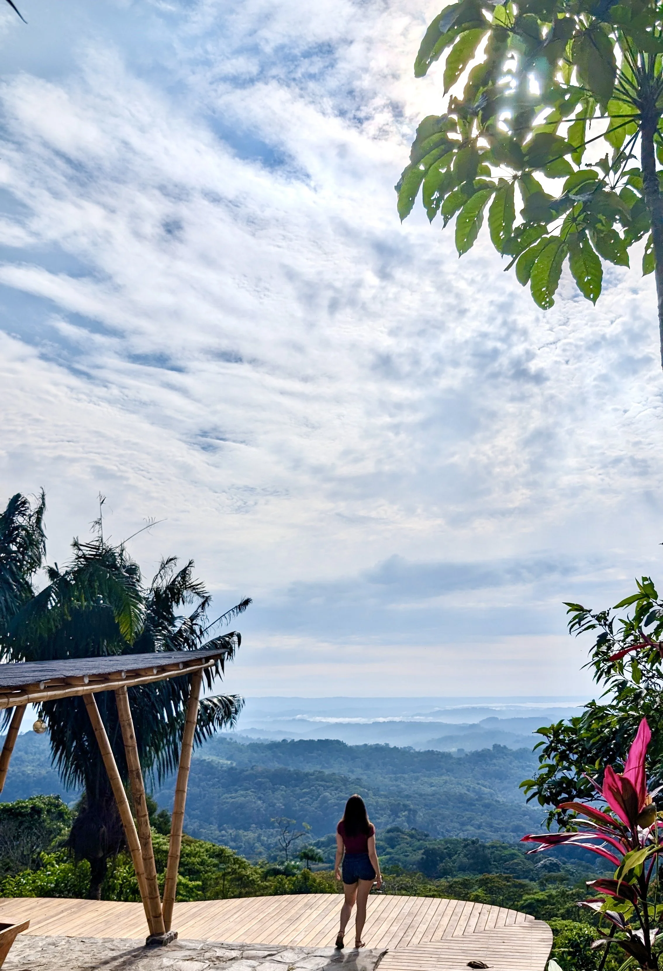 Una mujer de espaldas observa un paisaje de montañas y selva desde una plataforma de madera en un entorno natural con árboles y cielo nublado.