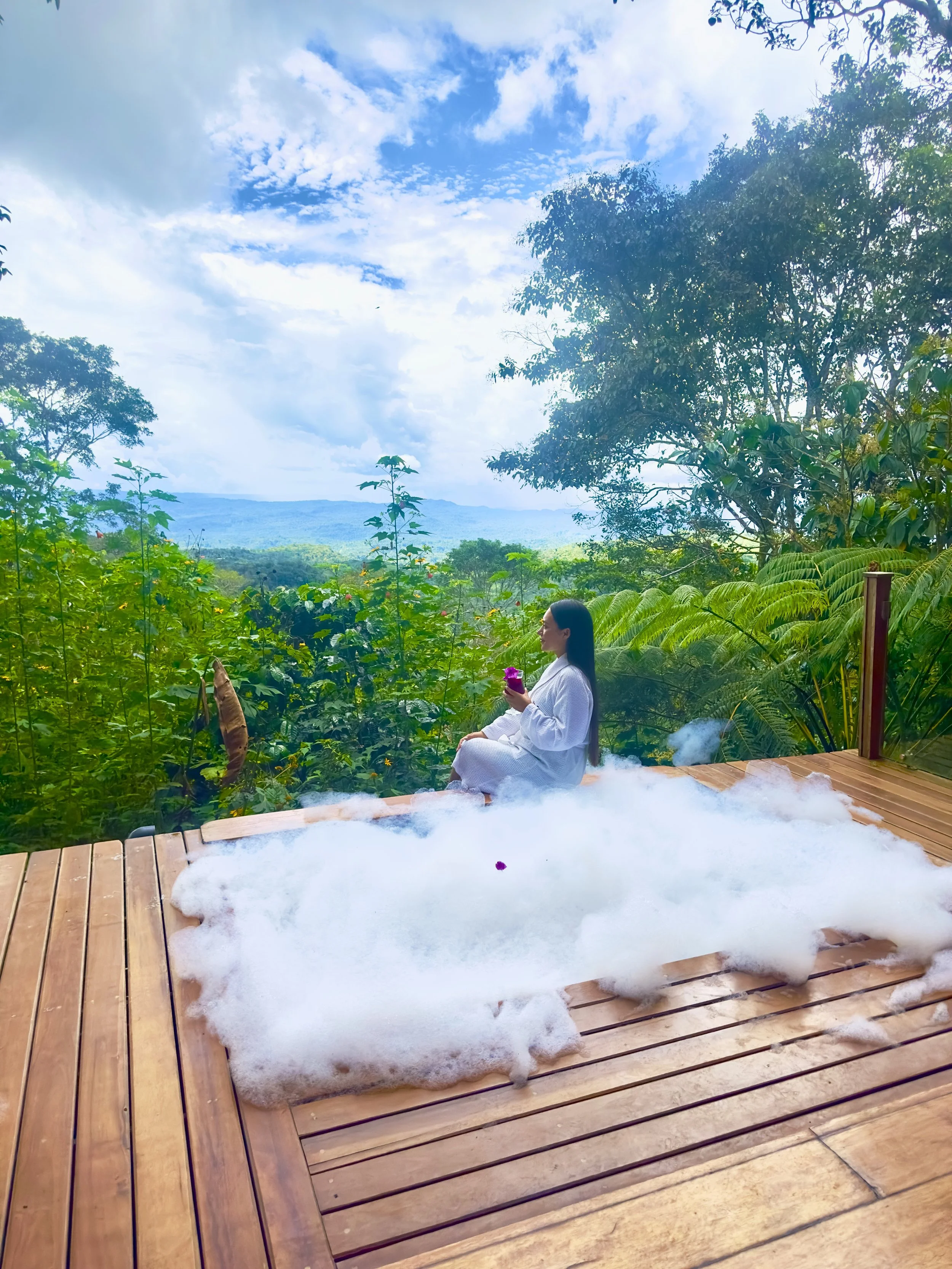 Mujer sentada en una terraza de madera con mucho vapor sobre ella, rodeada de vegetación y con un paisaje de montañas y cielo lluvioso al fondo.