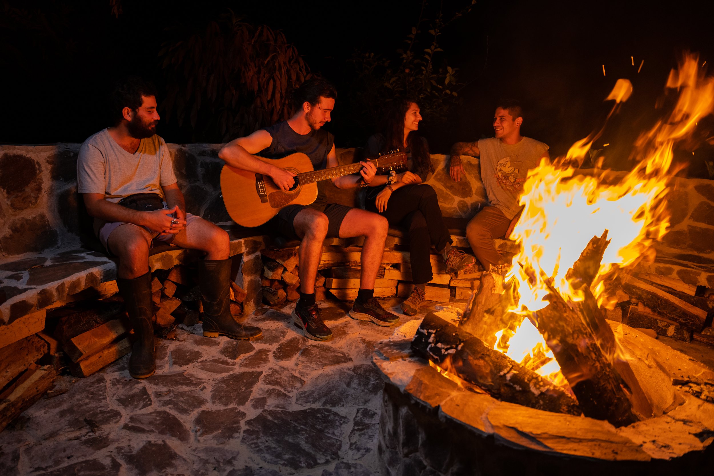 Grupo de cinco personas alrededor de una fogata en la noche, una tocando guitarra y las demás conversando y sonriendo.
