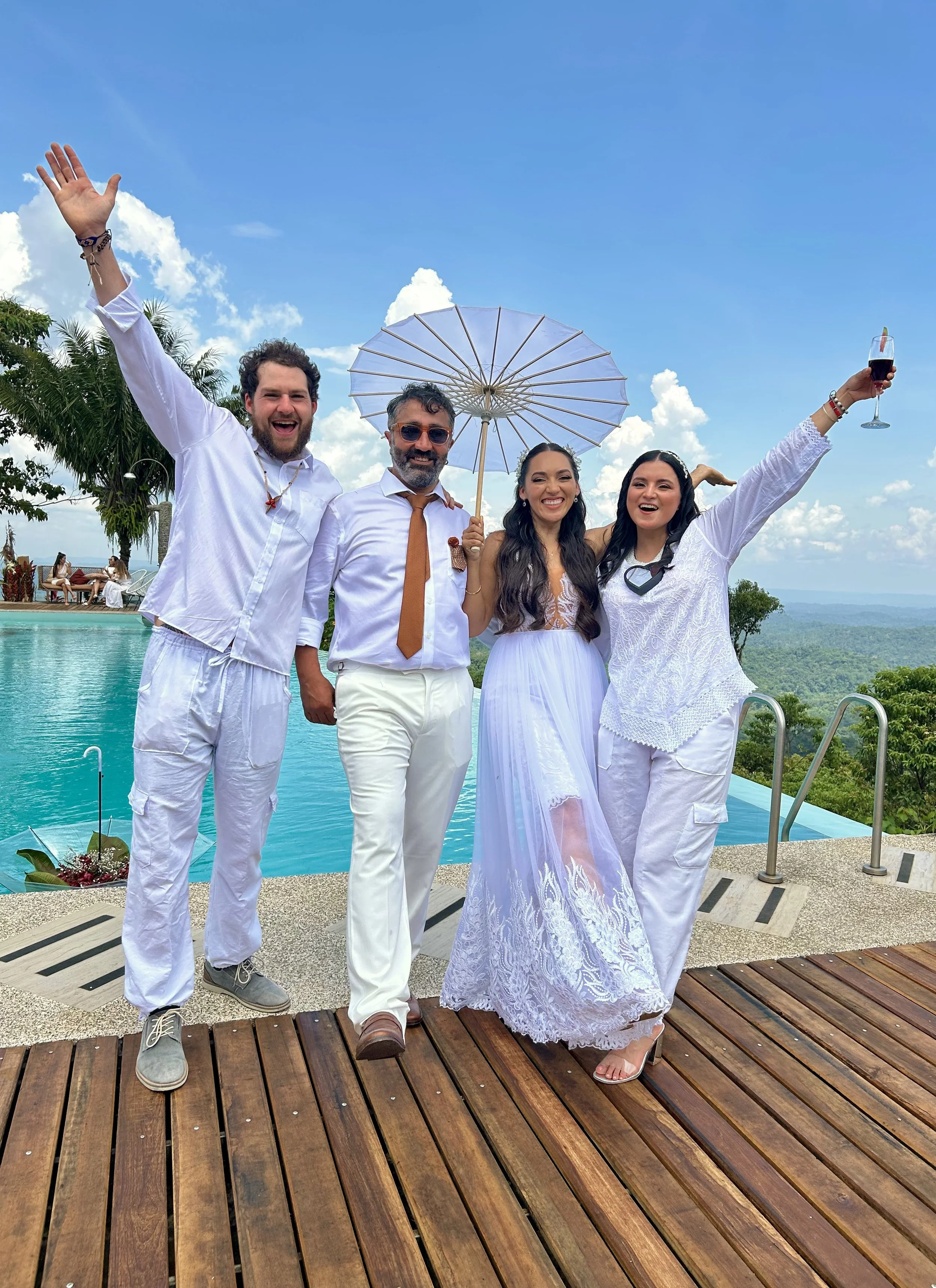 Grupo de cinco personas en una celebración cerca de la alberca, con fondo de paisaje montañoso y cielo azul con nubes, vestido de blanco y con sombrilla. Algunos con copas de vino.