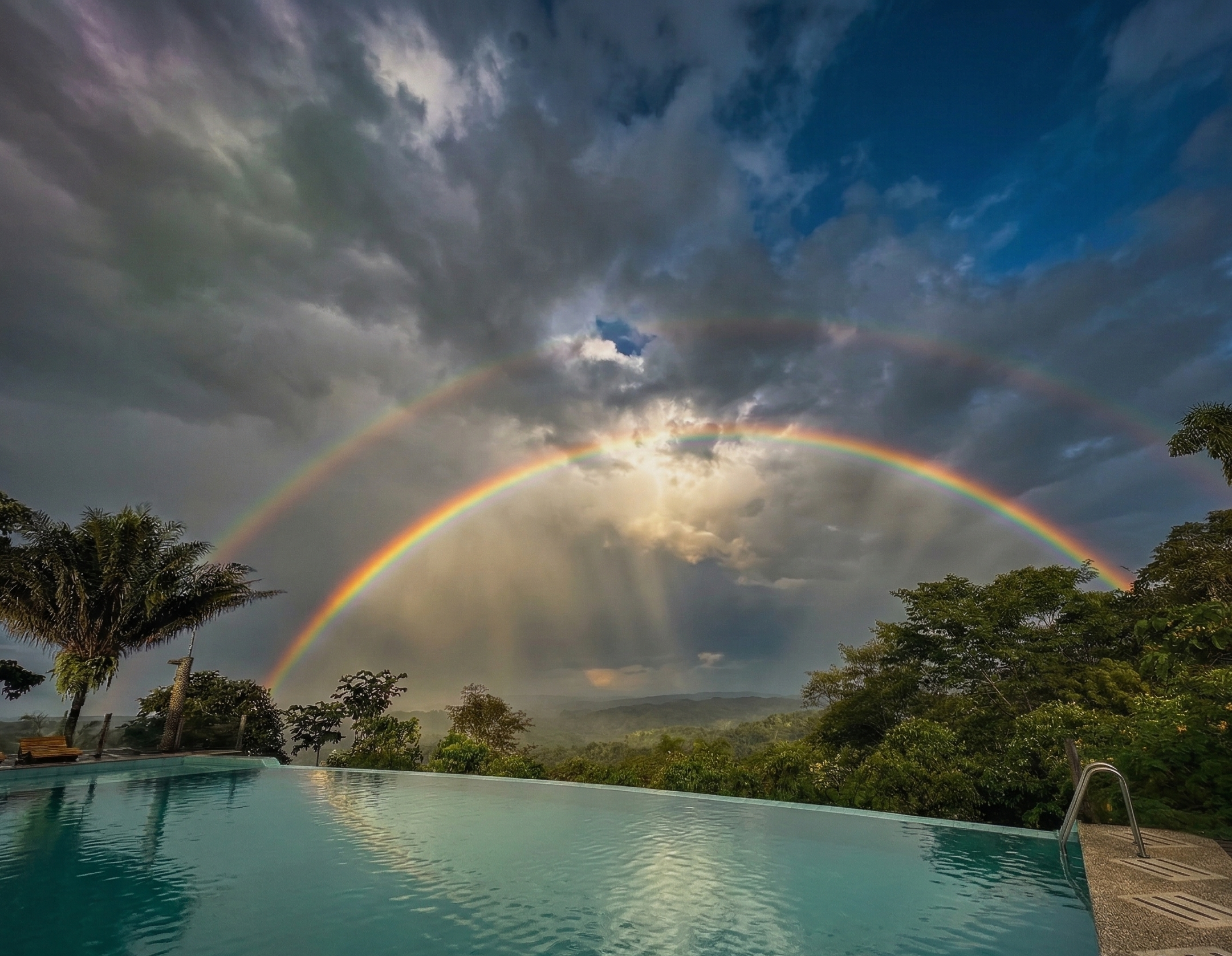 Piscina con árboles y un arcoíris doble en el cielo nublado, vista panorámica del paisaje montañoso.
