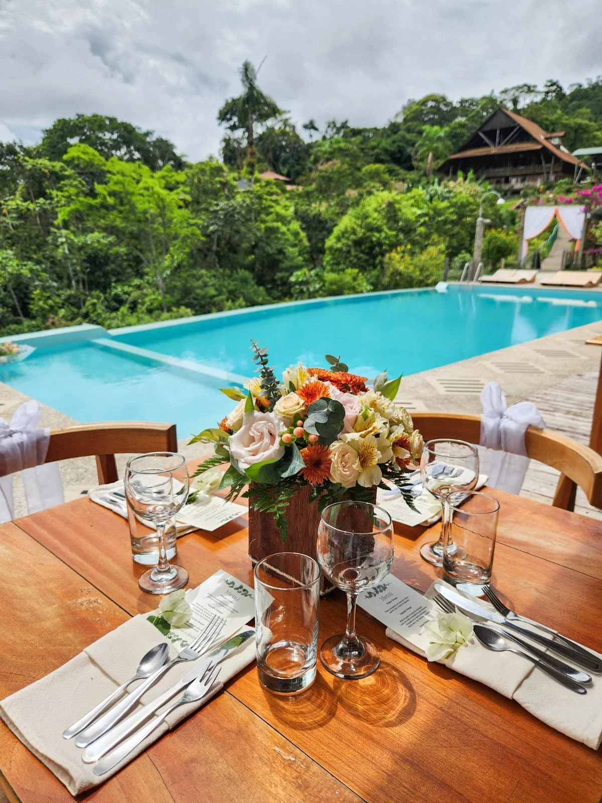 Mesa de comedor decorada con flores y utensilios, vista a una piscina y vegetación en un entorno natural.
