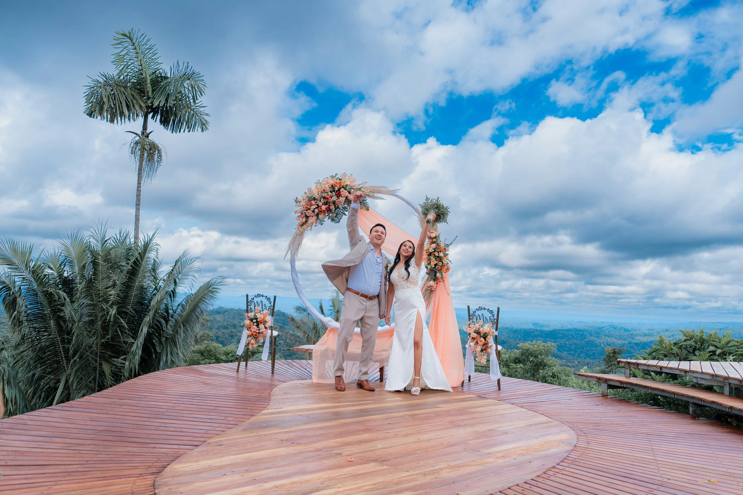 Pareja de recién casados en un escenario al aire libre con vista panorámica de montañas y cielos con nubes, en un decorado con flores y un arco de boda.
