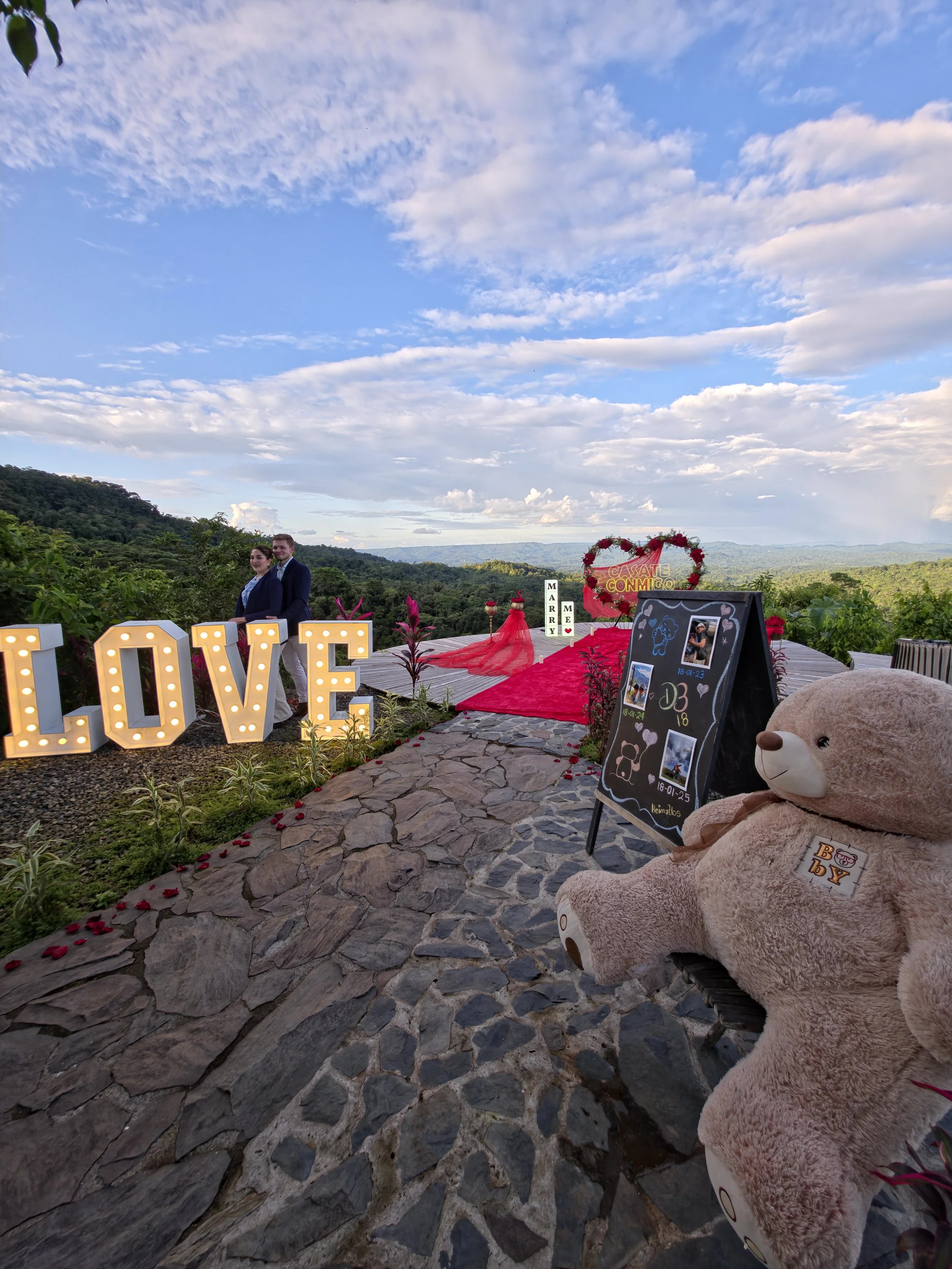 Escenario al aire libre con decoraciones para boda, including letras gigantes que dicen 'LOVE', una pareja en el fondo y un oso de peluche grande en primer plano, con un paisaje montañoso y cielo despejado.