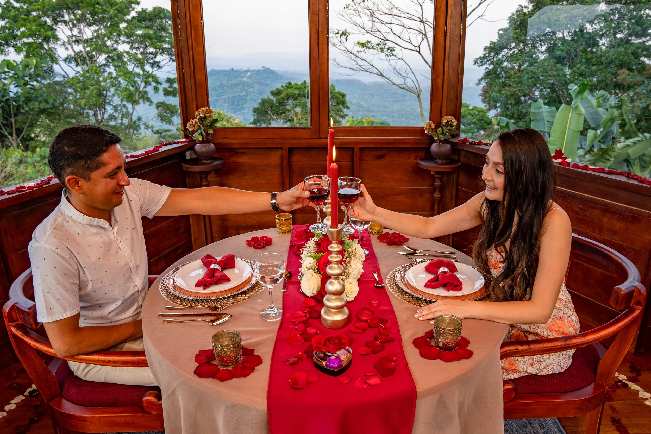 Pareja en una cena romántica en la torre Guacamayo con vista a la naturaleza, brindando con vino en una mesa decorada con flores, velas y pétalos de rosa.