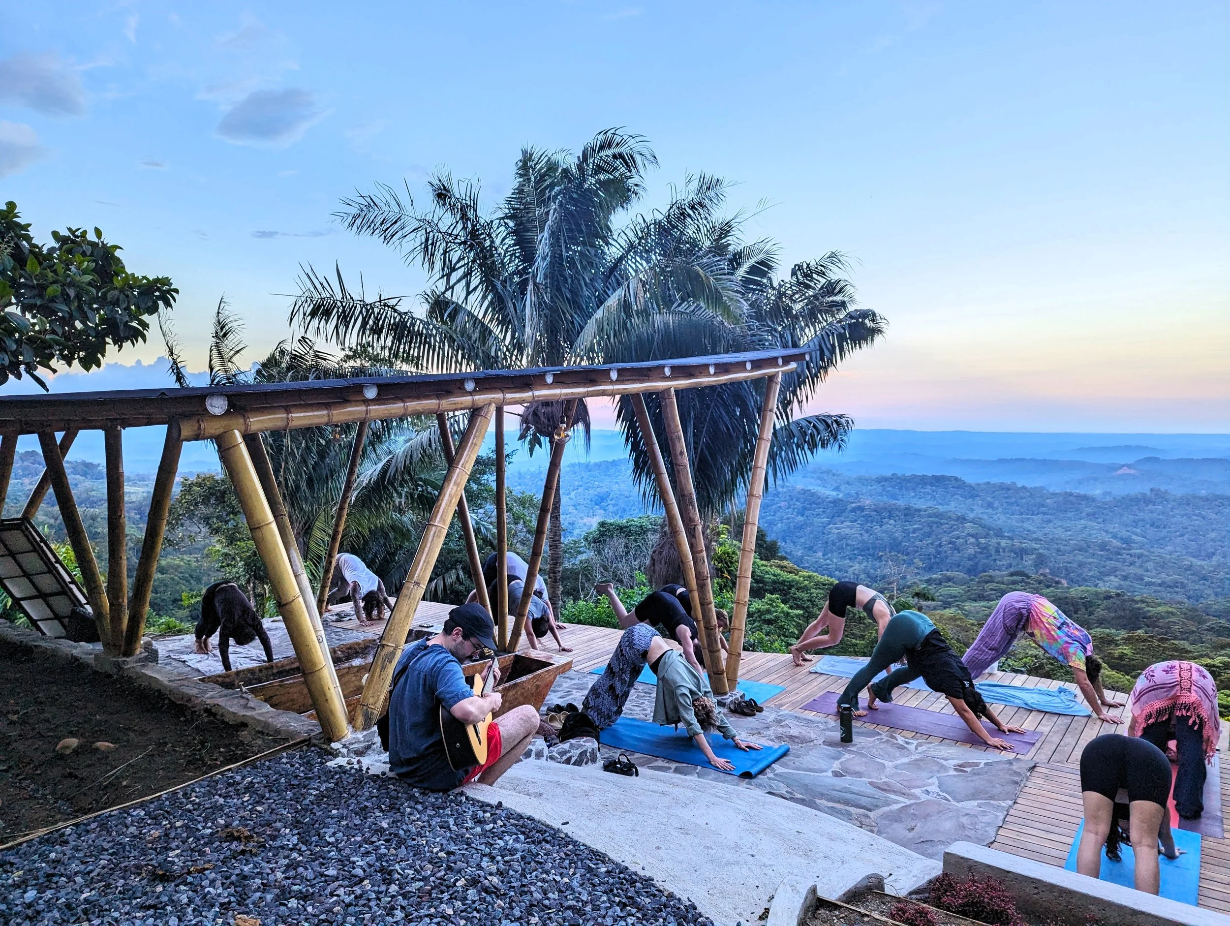 Grupo de personas haciendo yoga en el mirador con vista a un paisaje montañoso y selva al atardecer, con un instructor tocando guitarra.