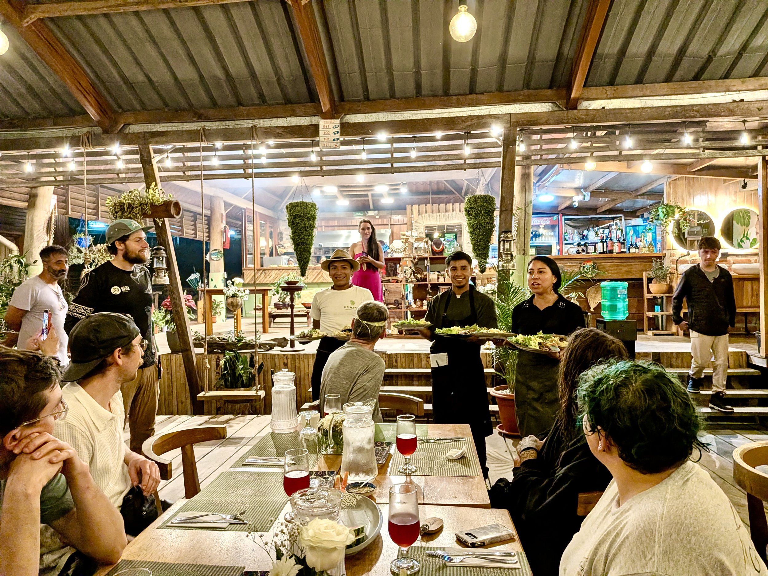 Personas en un restaurante observando a los camareros que llevan platos con comida, ambiente acogedor y decorado con plantas.