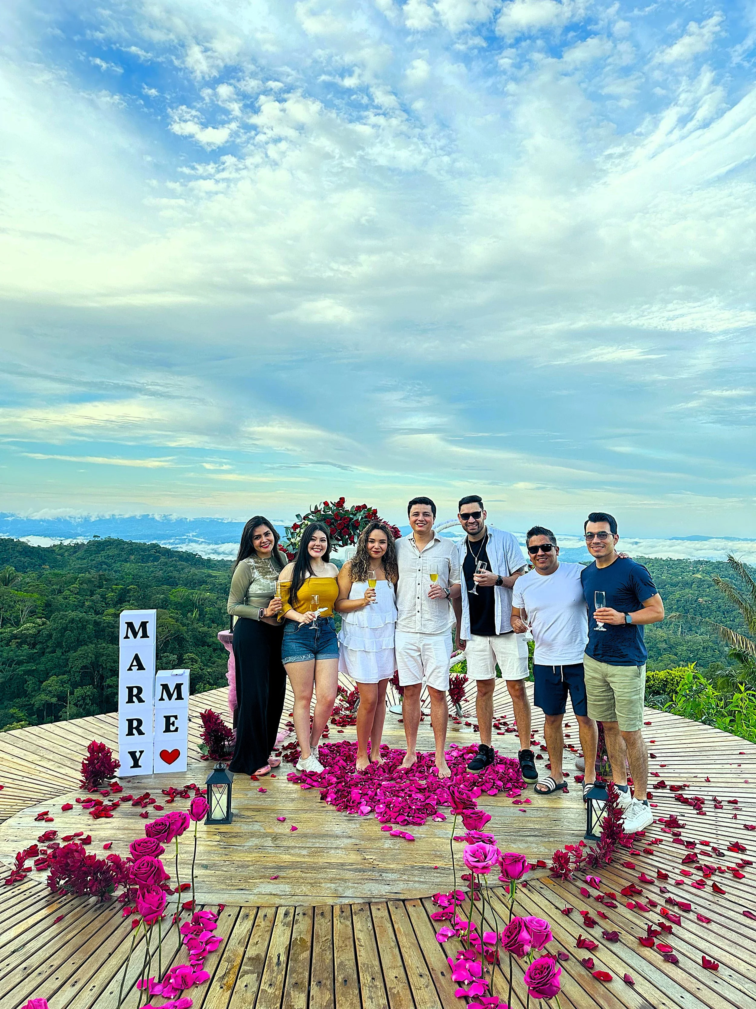 Grupo de siete personas en una pedida de mano al aire libre con cielo despejado y paisaje montañoso al fondo, decorado con rosas rosadas y letras que dicen 'Marry Me'.