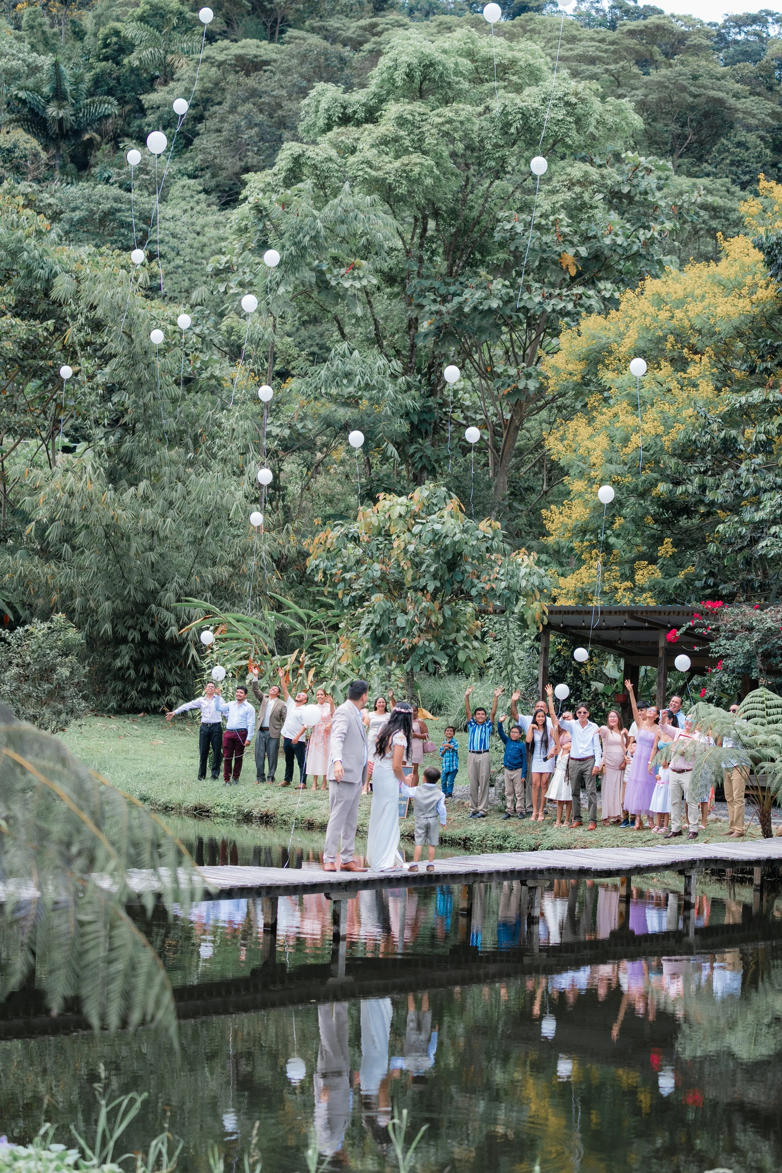 Grupo de personas en una celebración junto a un lago, lanzando globos blancos al aire, rodeados de árboles verdes