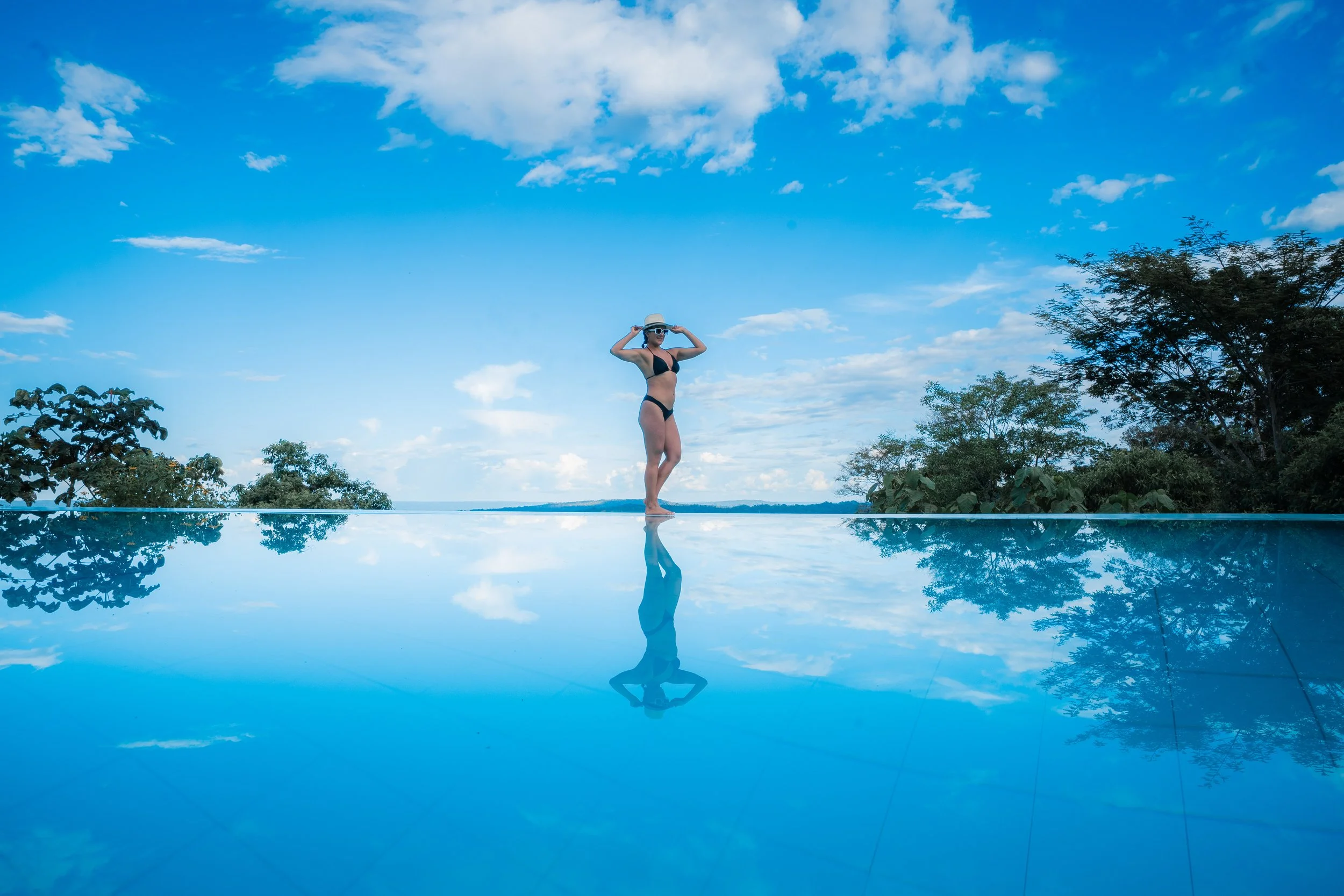 Mujer en traje de baño con sombrero y gafas sobre un borde de piscina con reflejo en el agua, árboles y cielo azul con nubes al fondo.