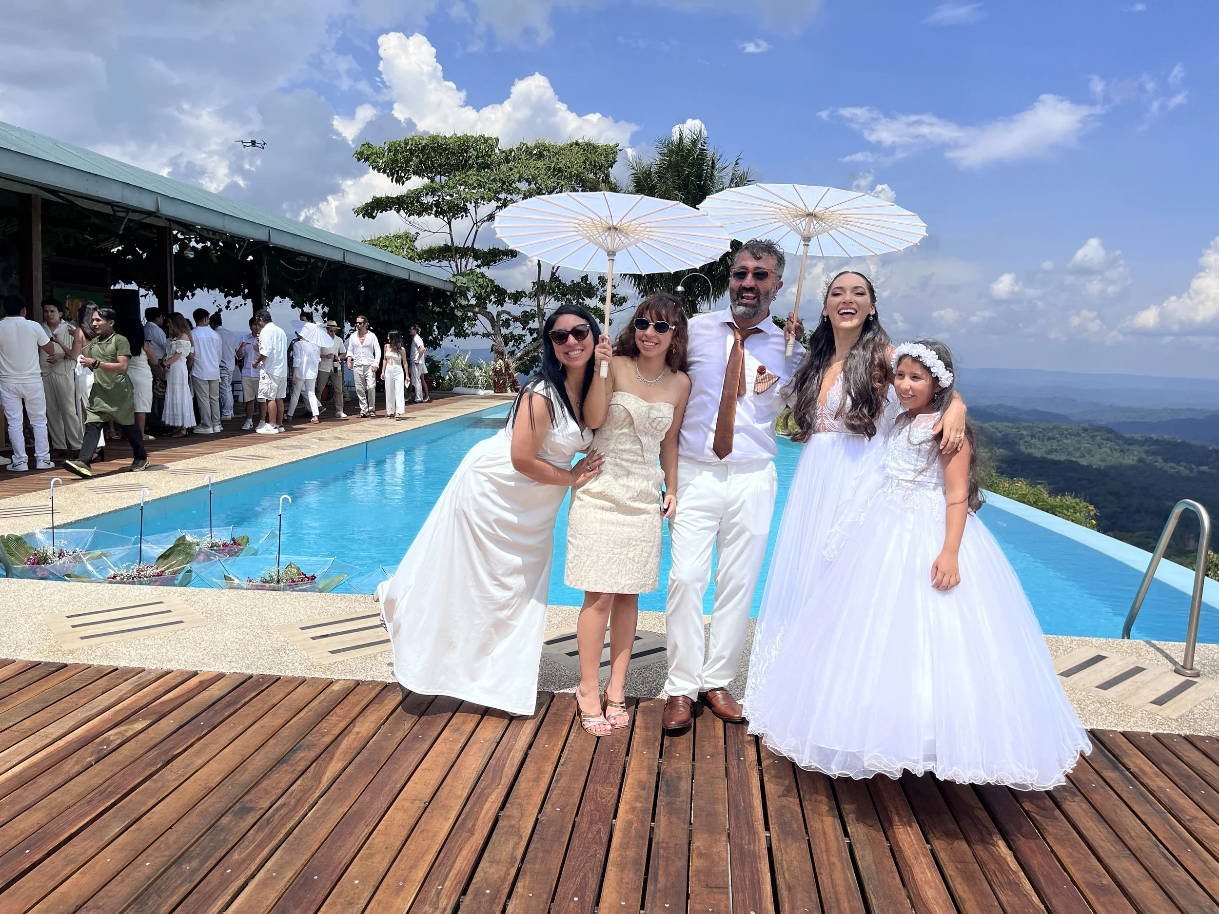Grupo de personas en una celebración junto a una piscina, con fondo de montañas y cielo azul, algunos con sombrillas y todos vestidos de blanco, con una boda o evento formal.