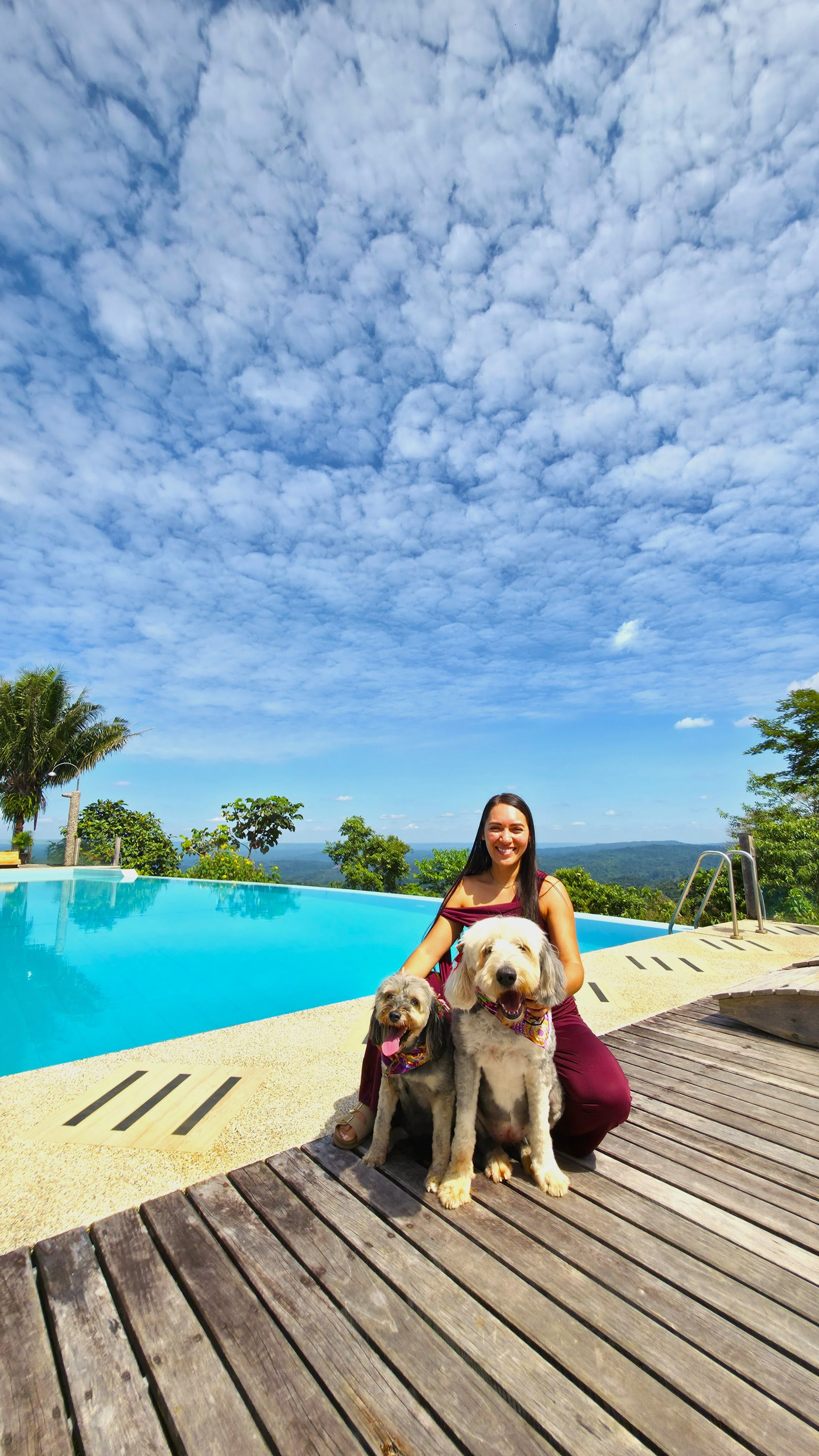 Una mujer con dos perros en una sala junto a una piscina con vistas a una zona de naturaleza llana y con árboles, en un día soleado con cielo parcialmente nublado.