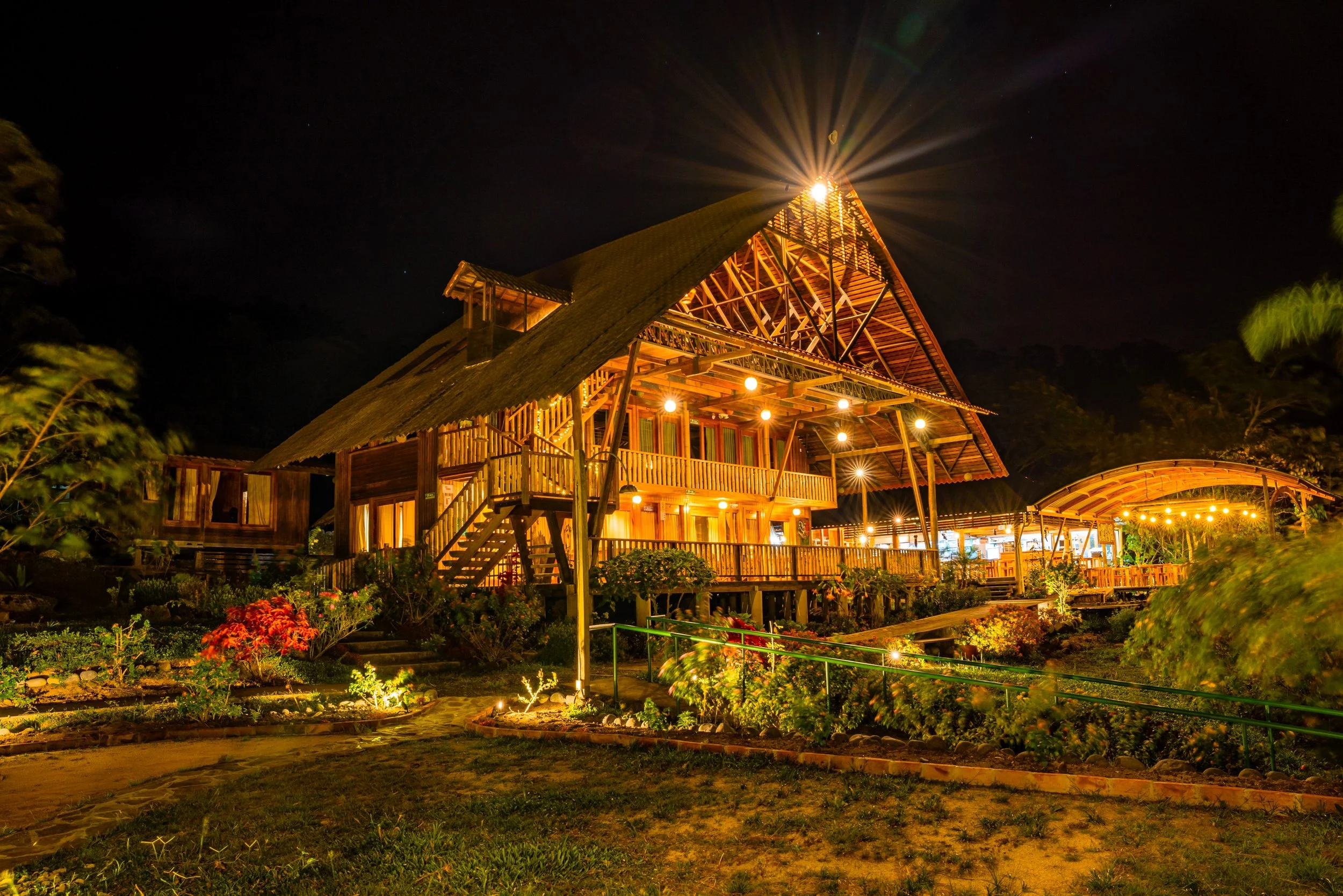 Casa Grande de madera iluminada con luces en la noche, rodeada de jardines y plantas.