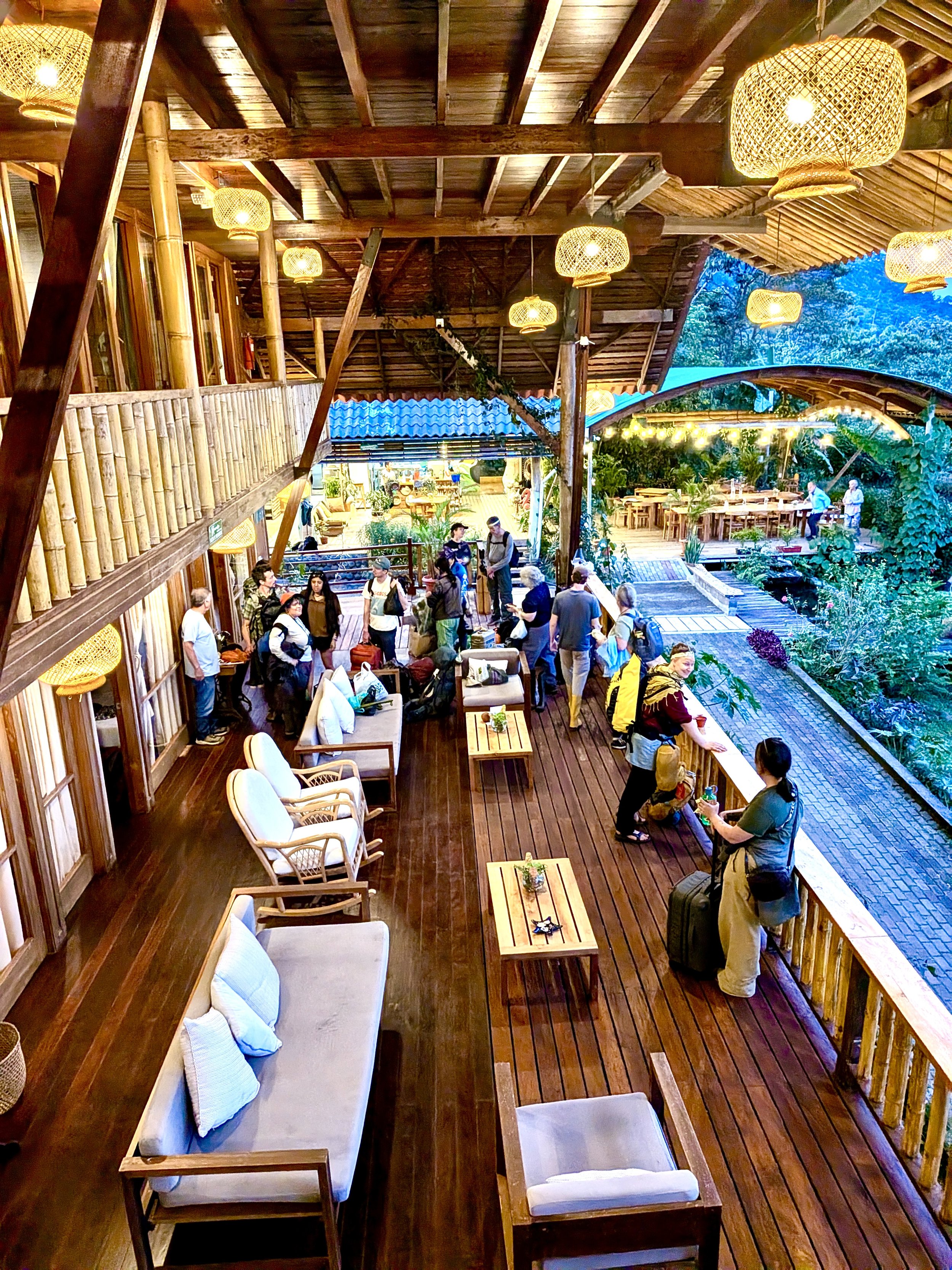 La terraza de la Casa Grande y decoración natural. Hay varias personas, algunas con maletas, y mesas ocupadas. La área exterior visible muestra un ambiente verde y relajante con iluminación cálida.