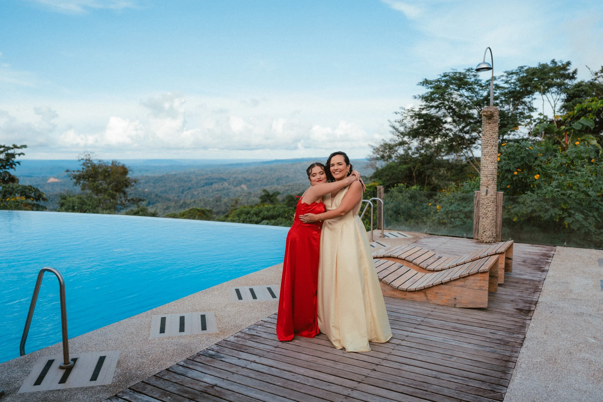 Mujeres abrazándose junto a una piscina con vista panorámica a un paisaje verde y celeste en un día soleado.