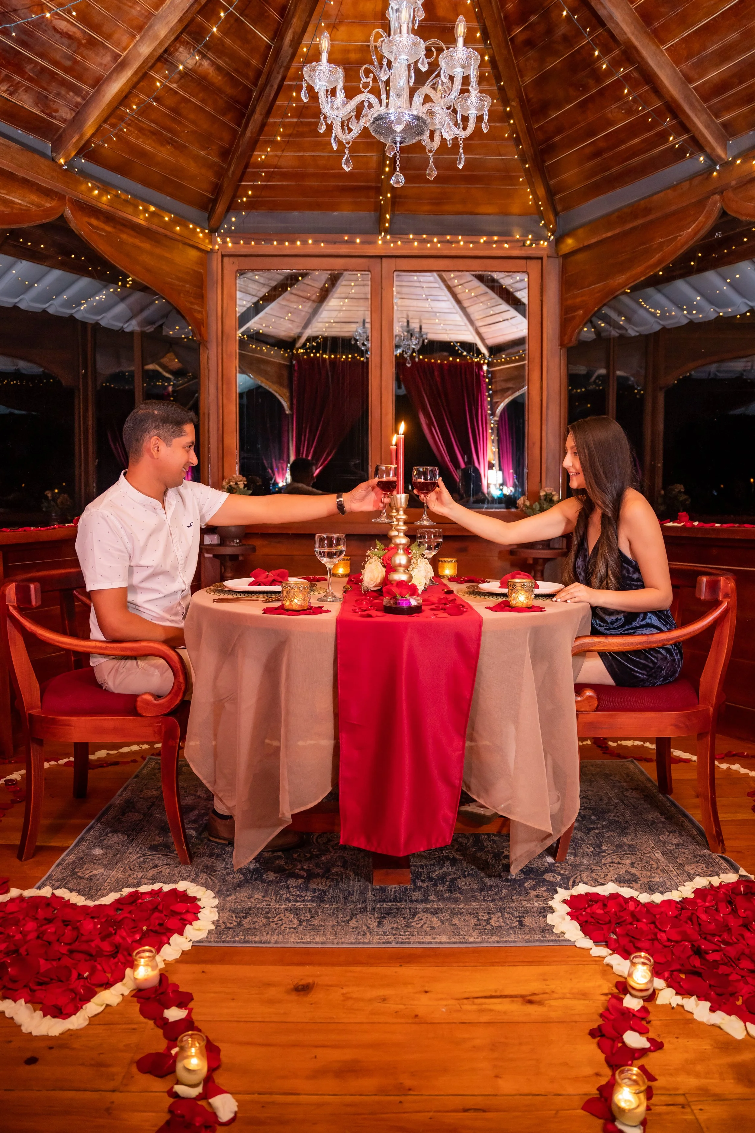 Una pareja celebra una cena romántica en un ambiente acogedor y decorado con luces y velas, con un centro de mesa con velas y flores, en un restaurante o mirador con paredes de madera y ventanas grandes.