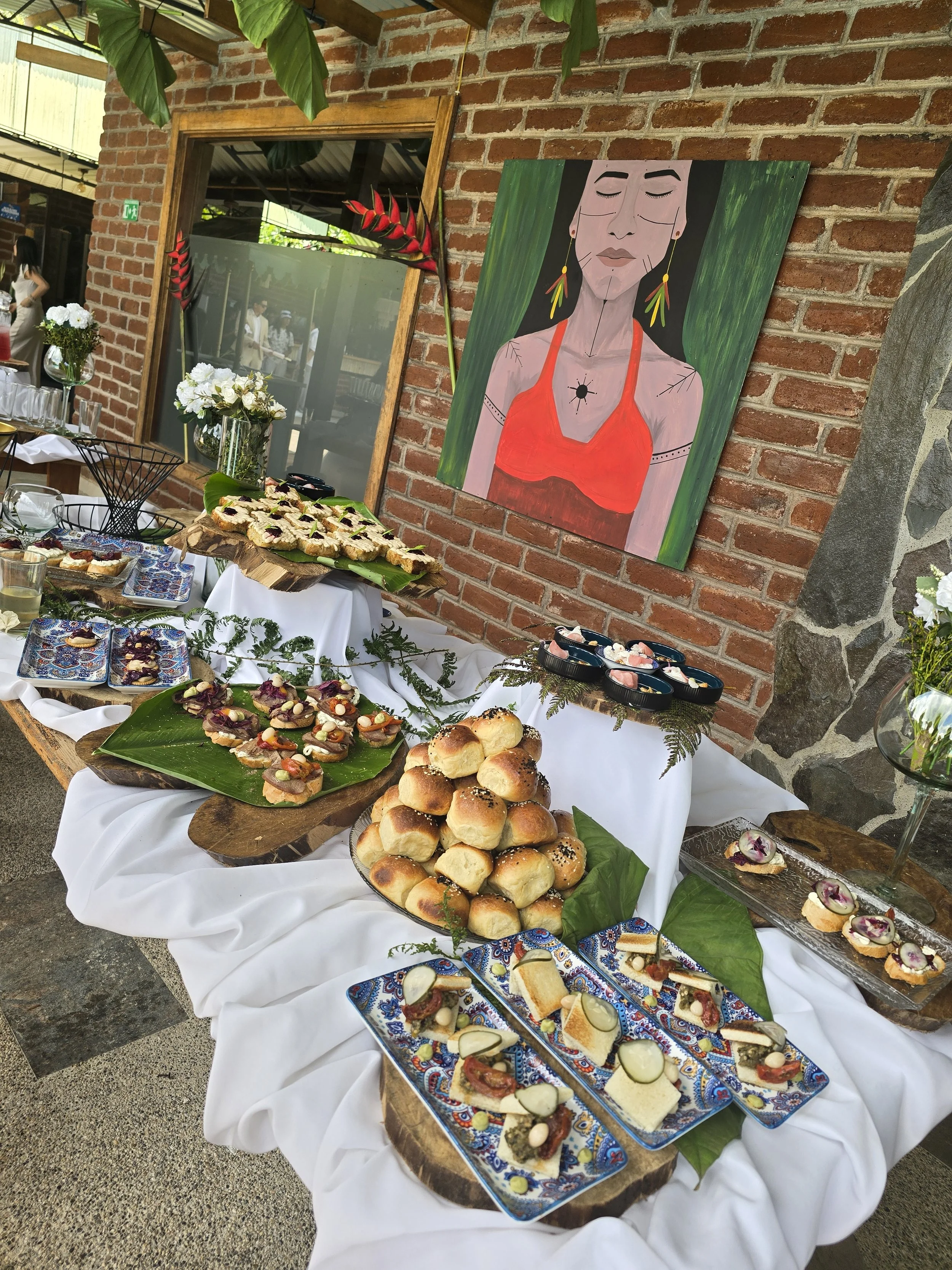 Mesa decorada con variedad de panes y bocadillos, adornada con hojas verdes y flores, en un ambiente rústico con pared de ladrillos y pintura de una mujer con pendientes, en un evento o celebración.