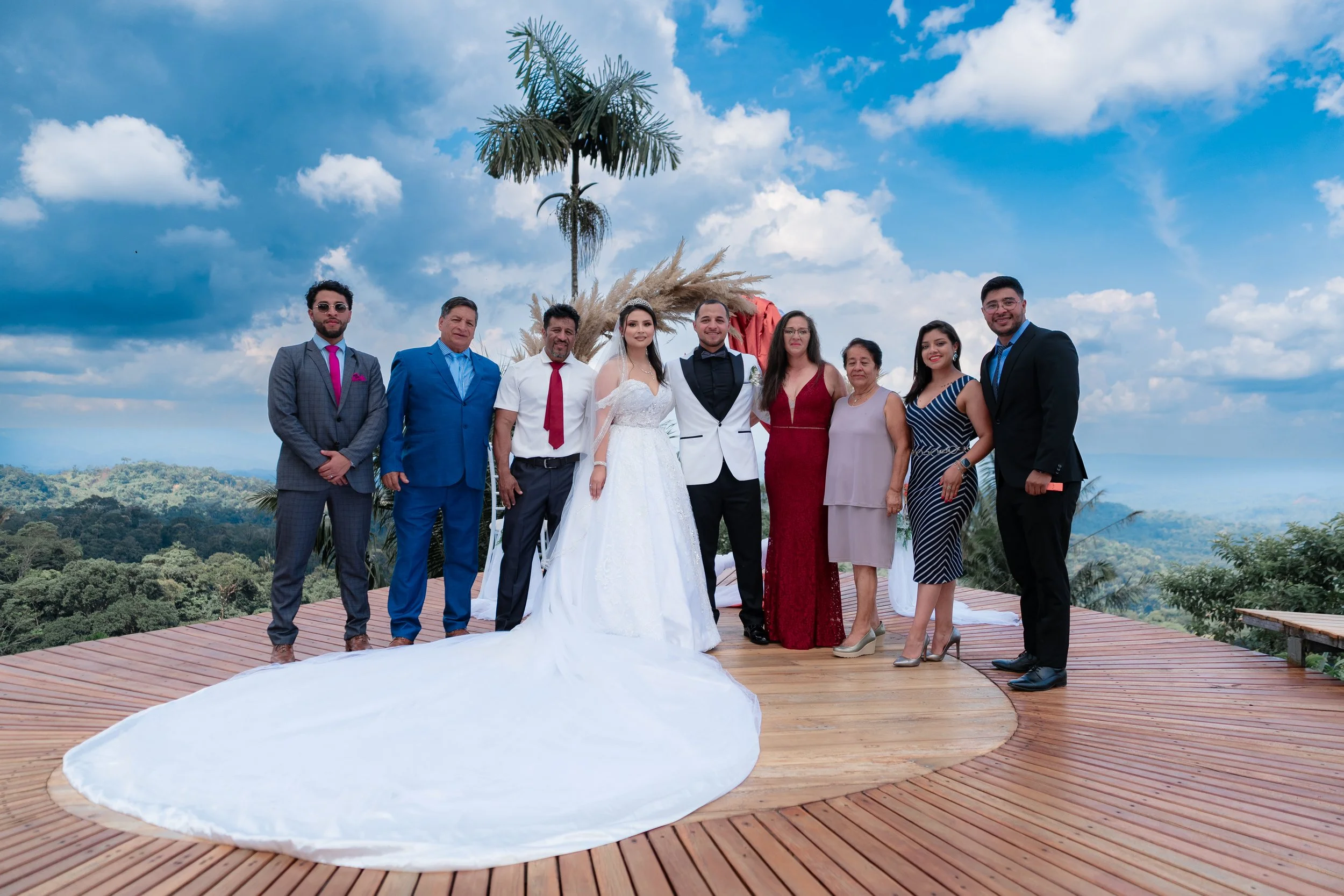 Grupo de personas en una ceremonia de matrimonio al aire libre, con fondo de cielo y naturaleza, en un escenario de madera decorado con palmeras y flores.
