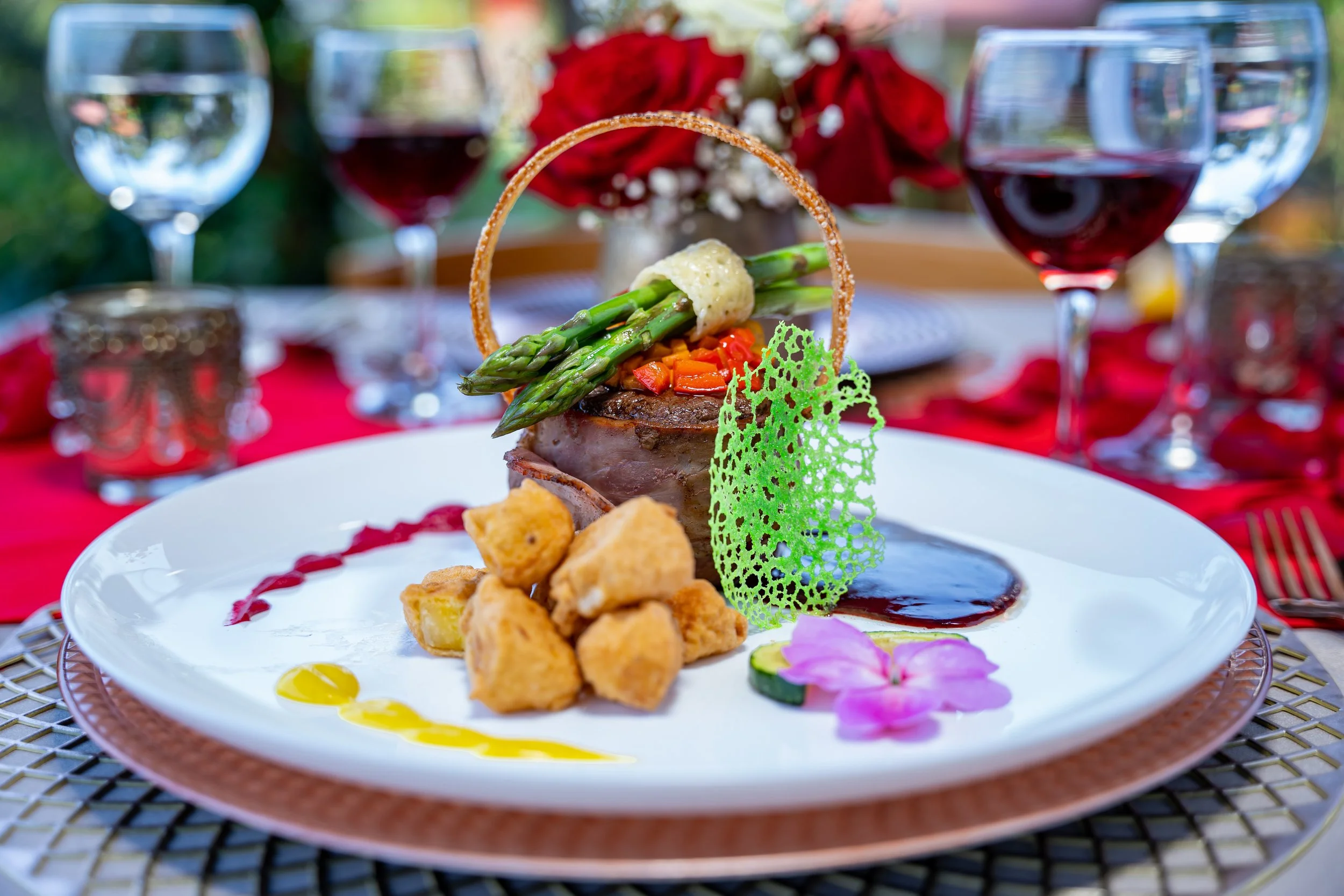 Plato gourmet de carne con verduras, patatas fritas, salsa y decoración floral en la mesa con copas de vino tinto.