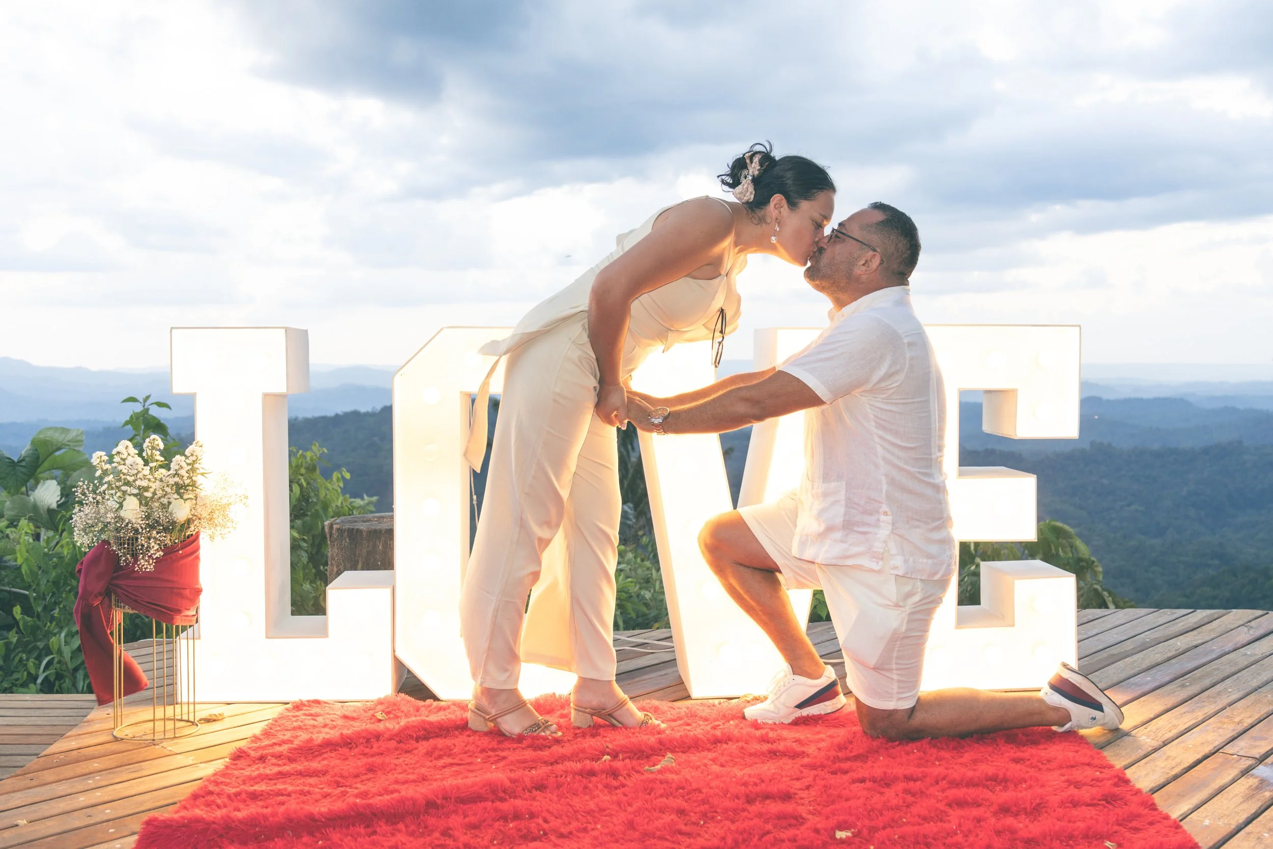 Pareja en ceremonia de propuesta de matrimonio: mujer besando a hombre en rodillas, con letras grandes que dicen 'LOVE' y un fondo de naturaleza y cielo.