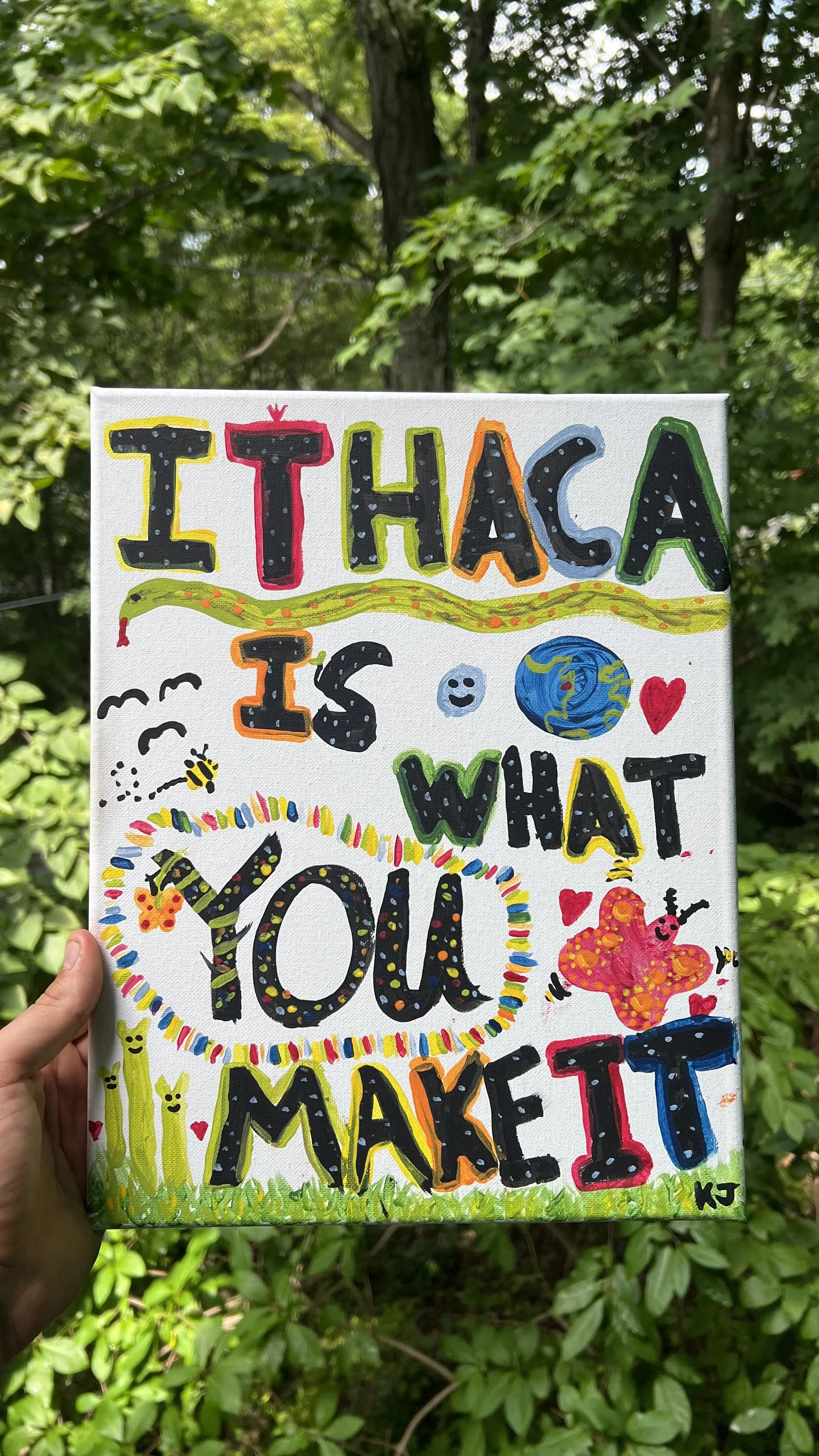Colorful hand-painted sign held in front of green trees with the message, 'I THOUGHT I WAS THE ONE WHO MADE IT,' decorated with hearts, a butterfly, a globe, and expressive doodles.