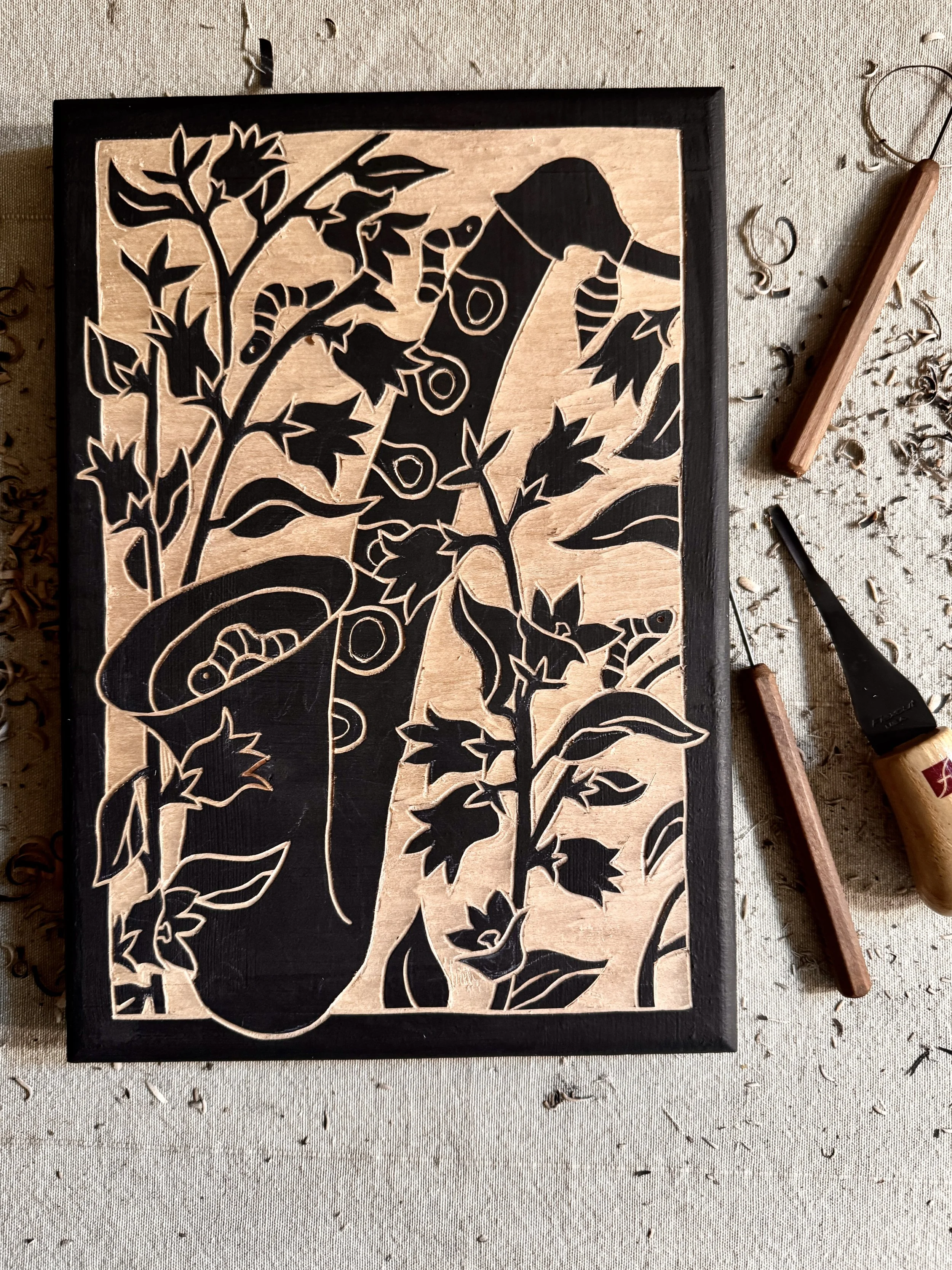 A wooden carving of a rabbit and garden scene carved into a rectangular piece of wood, placed on a work surface surrounded by carving tools, wood shavings, and scraps.