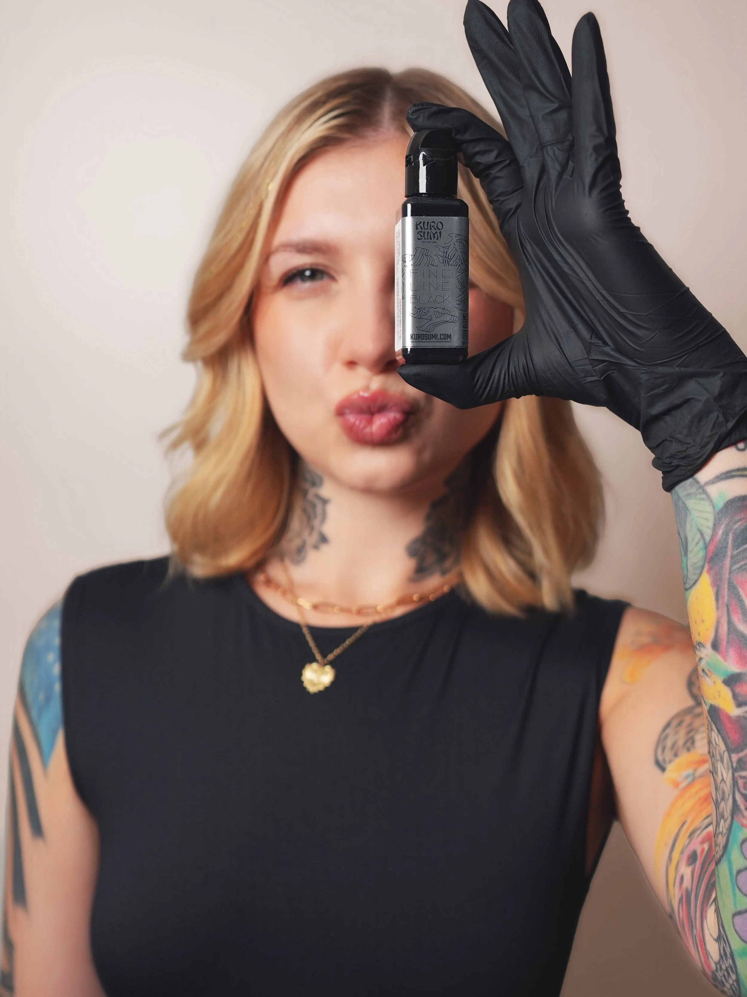 A young woman with shoulder-length brown hair is sitting on a cream-colored chair with gold accents in a cozy indoor space. She is wearing a blue sweatshirt that reads 'REVIVE BEAUTY BAR' and has a tattoo on her neck. Behind her, there is a small, frosted miniature Christmas tree on a side table, and wall decorations including a wooden shelf with mini trees, a neon pink sign that says 'love,' and gold snowflakes. There is also a beige tufted bench and a cup on the floor.