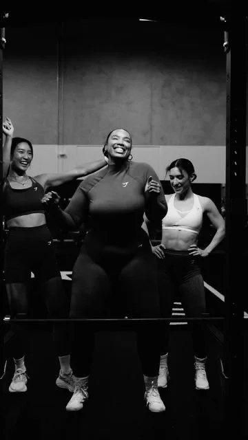Three women in workout clothes smiling, posing in front of a mirror in a gym.