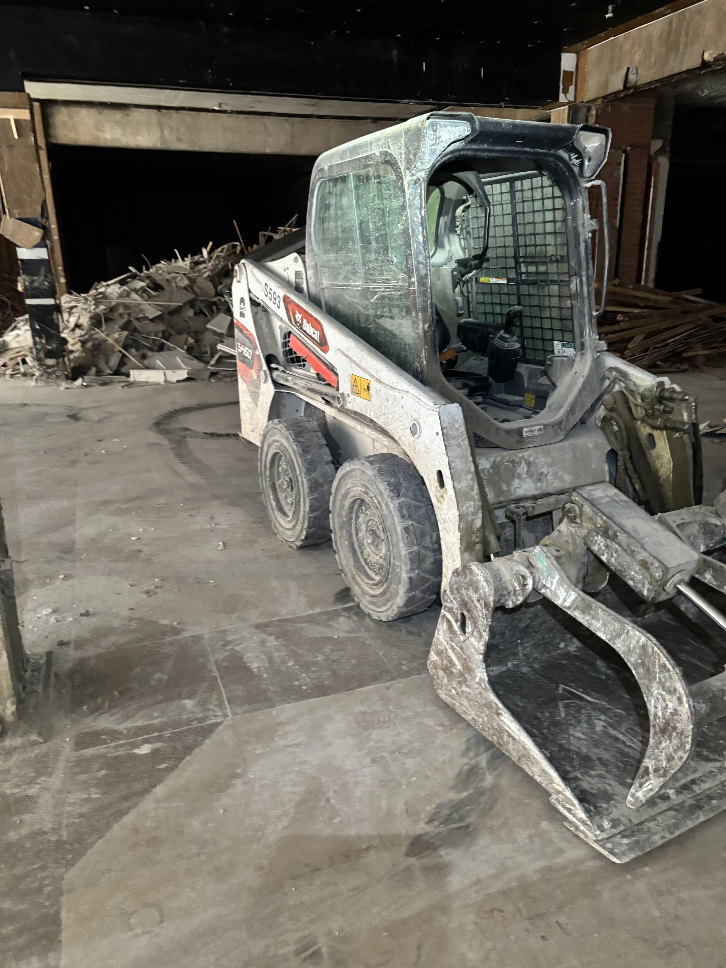 A construction site with a mini skid-steer loader and a pile of debris and wooden planks in the background, inside an unfinished building.
