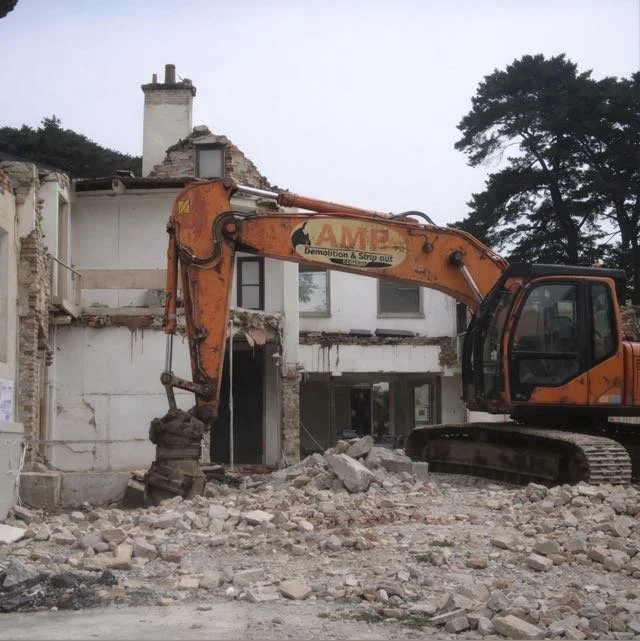 An orange excavator demolishing a partially destroyed building with rubble on the ground.