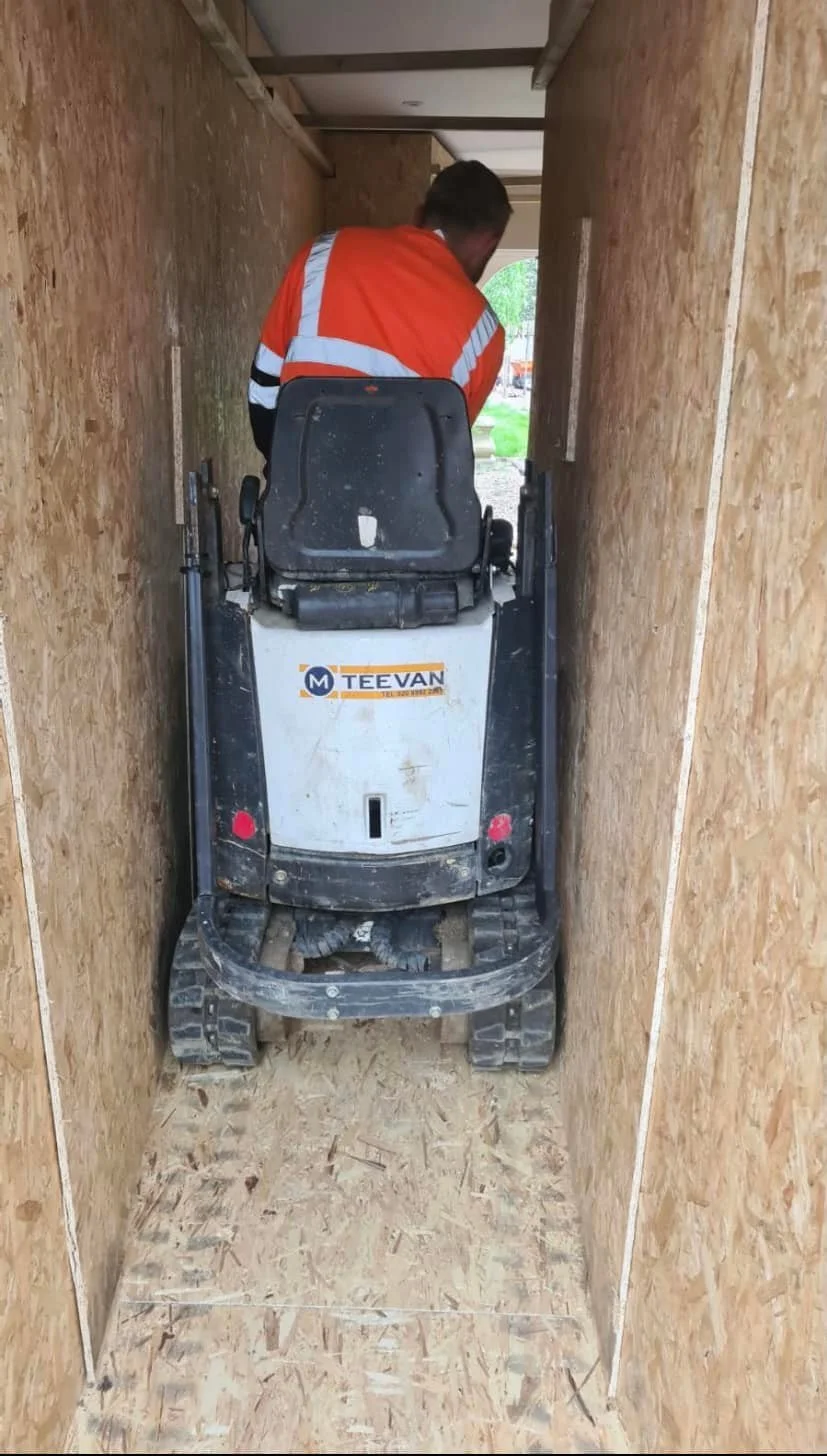 Construction worker in an orange safety vest operating a small tracked excavator in a narrow wooden hallway under construction.