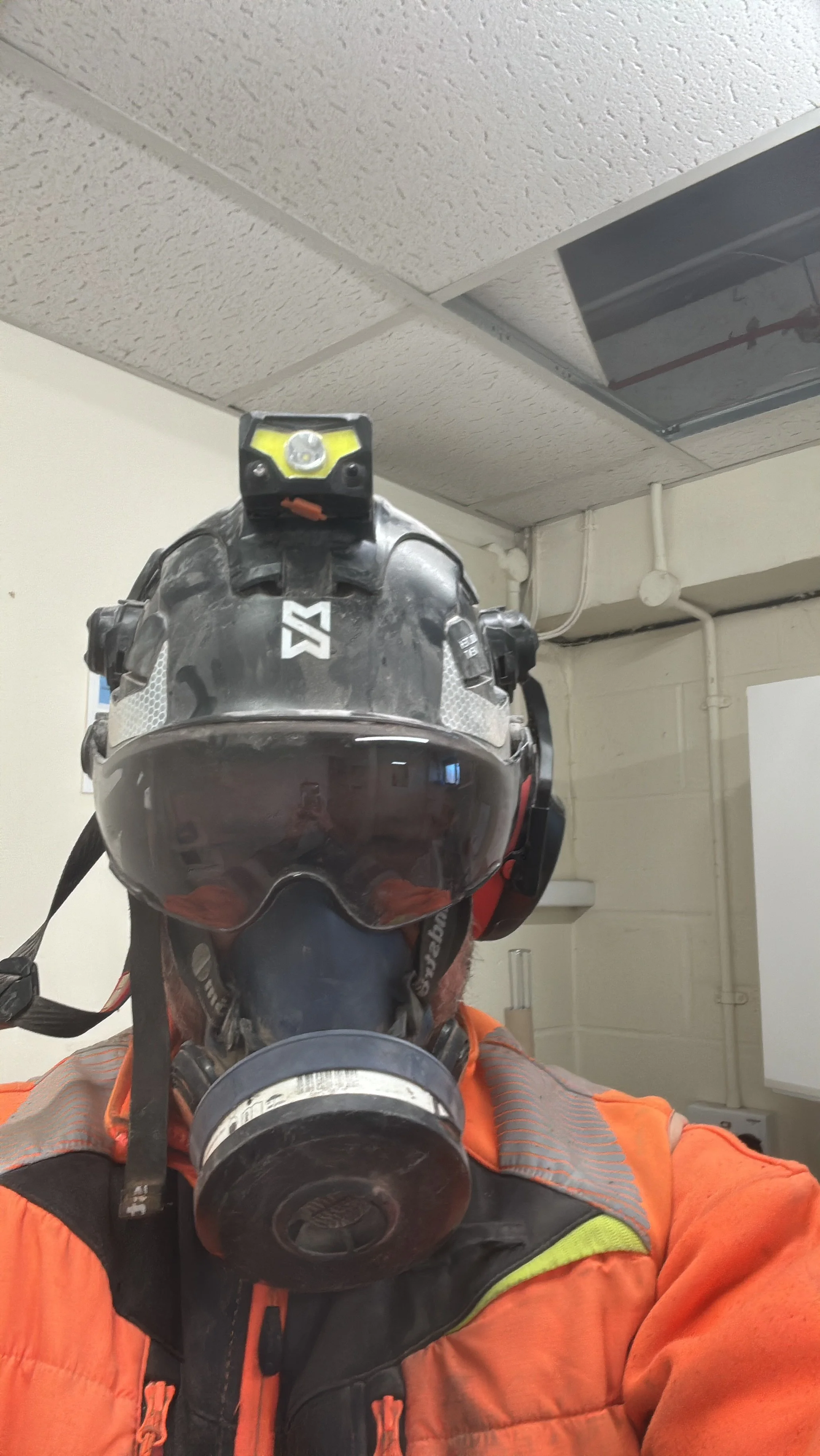 A person wearing a helmet with a mounted headlamp and an orange safety jacket, taking a selfie indoors.
