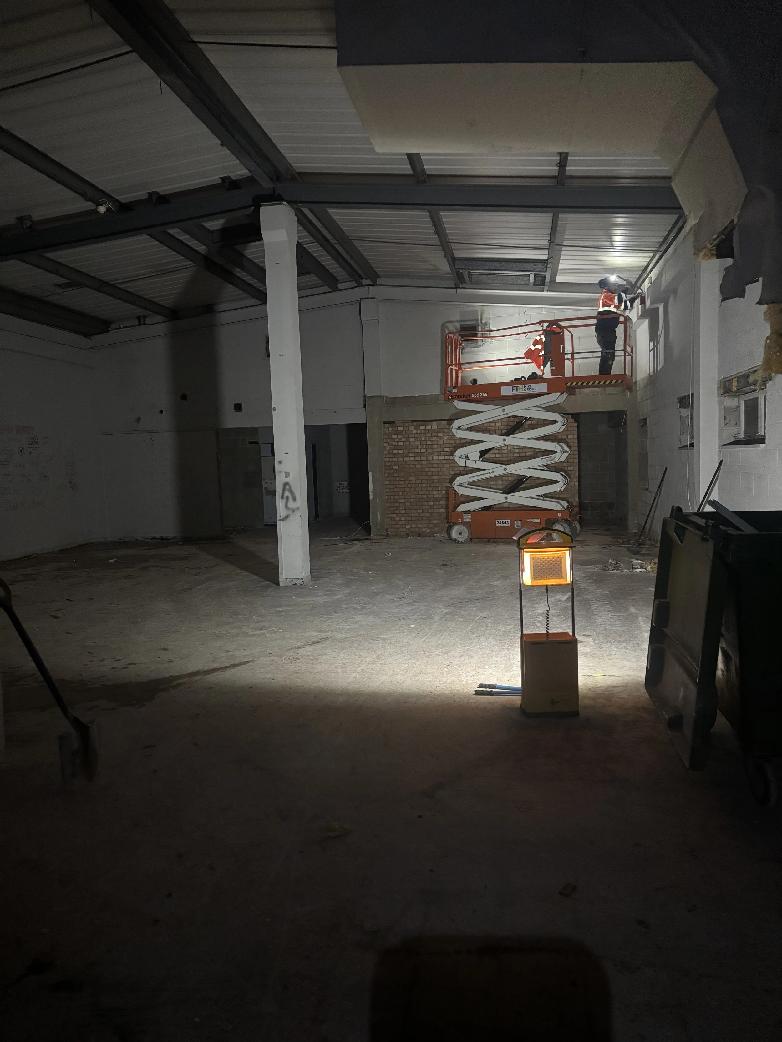 Construction workers on a scissor lift working on a wall in an unfinished building. The space is well lite in the work area. 