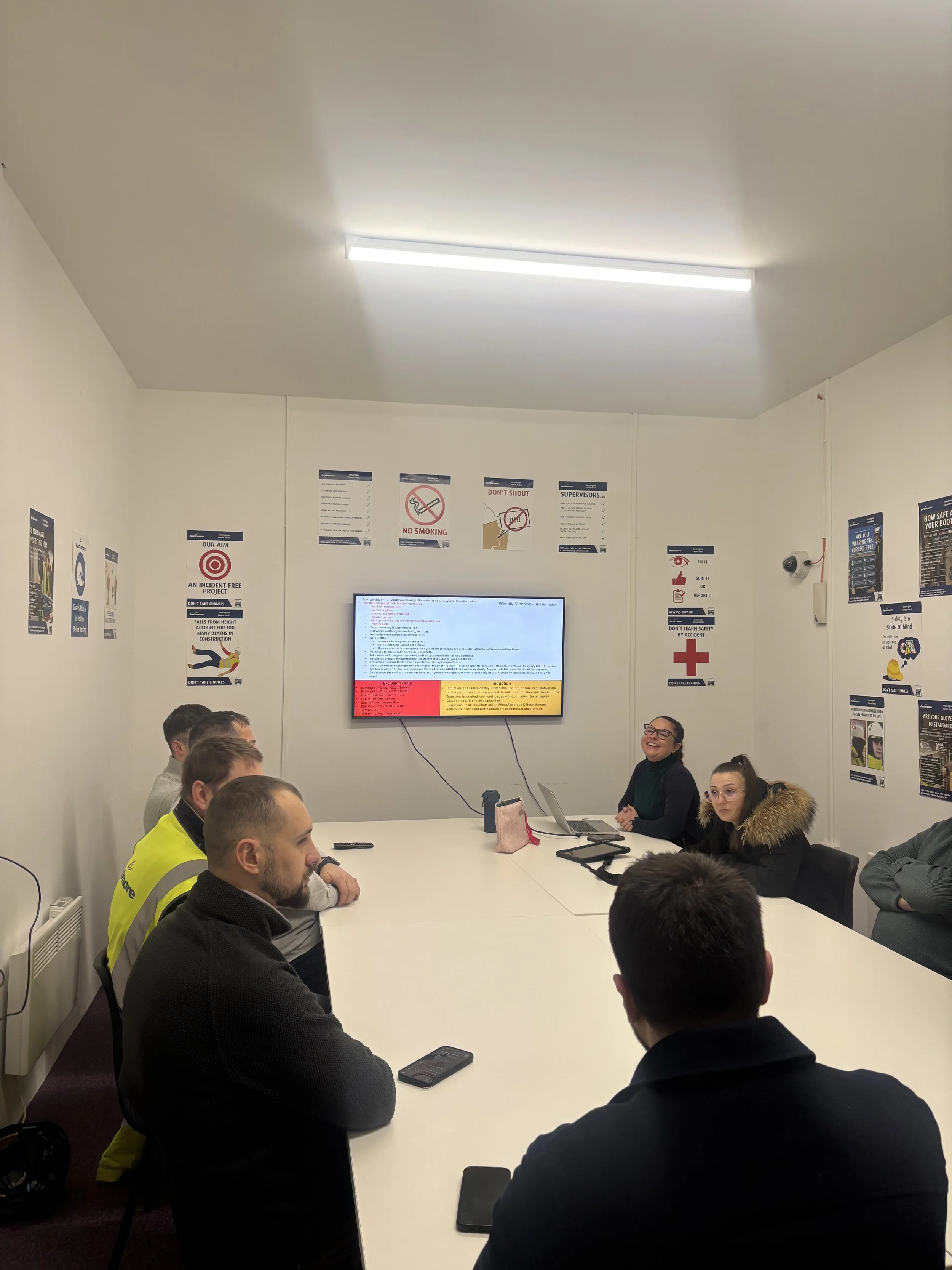 People sitting around a white conference table in a meeting room, listening to a presentation on a wall-mounted screen, with safety posters on the wall.