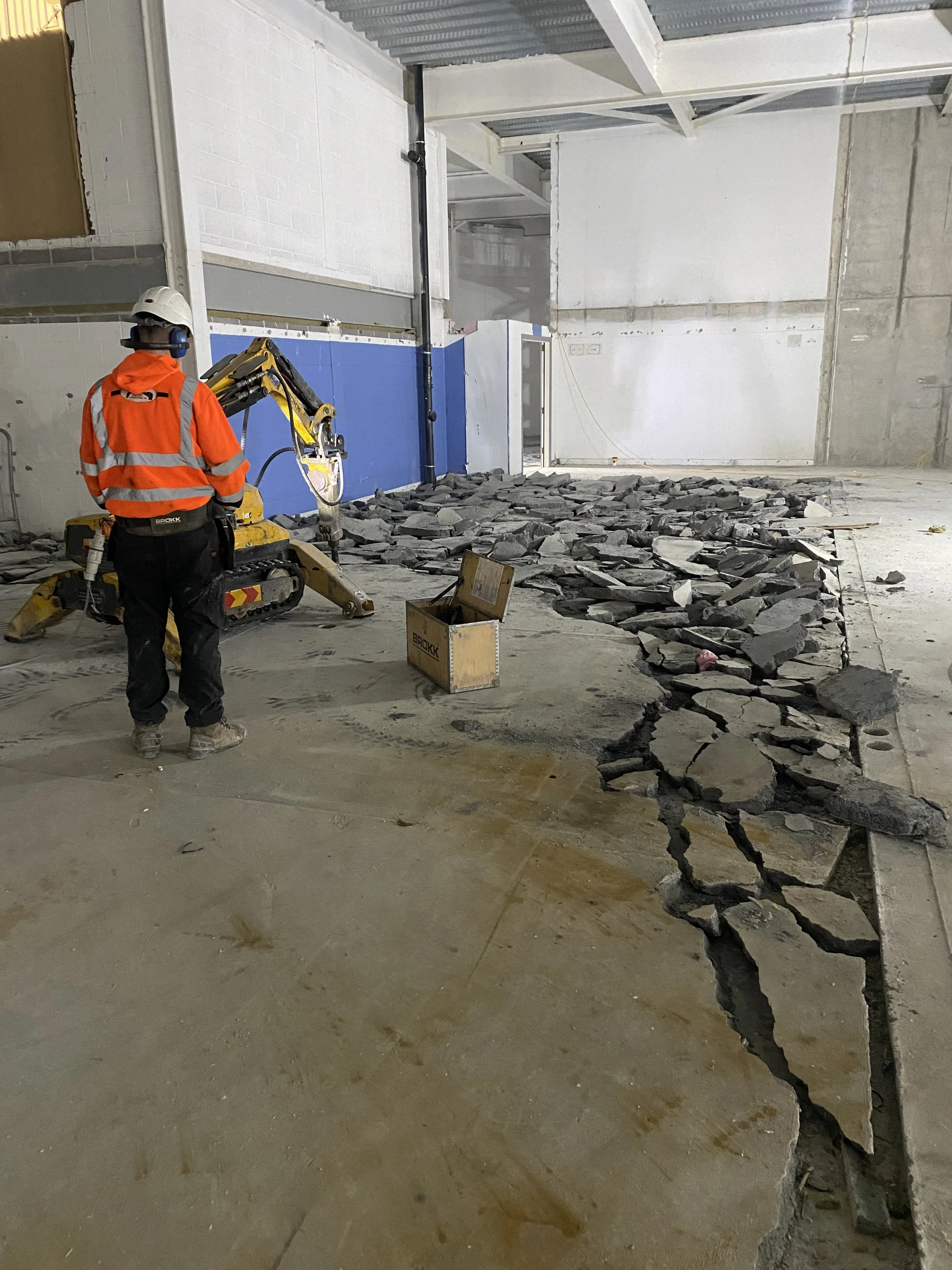 Construction worker wearing orange safety jacket and white helmet operating a small yellow excavator in an unfinished building with broken concrete on the floor and partially built walls.