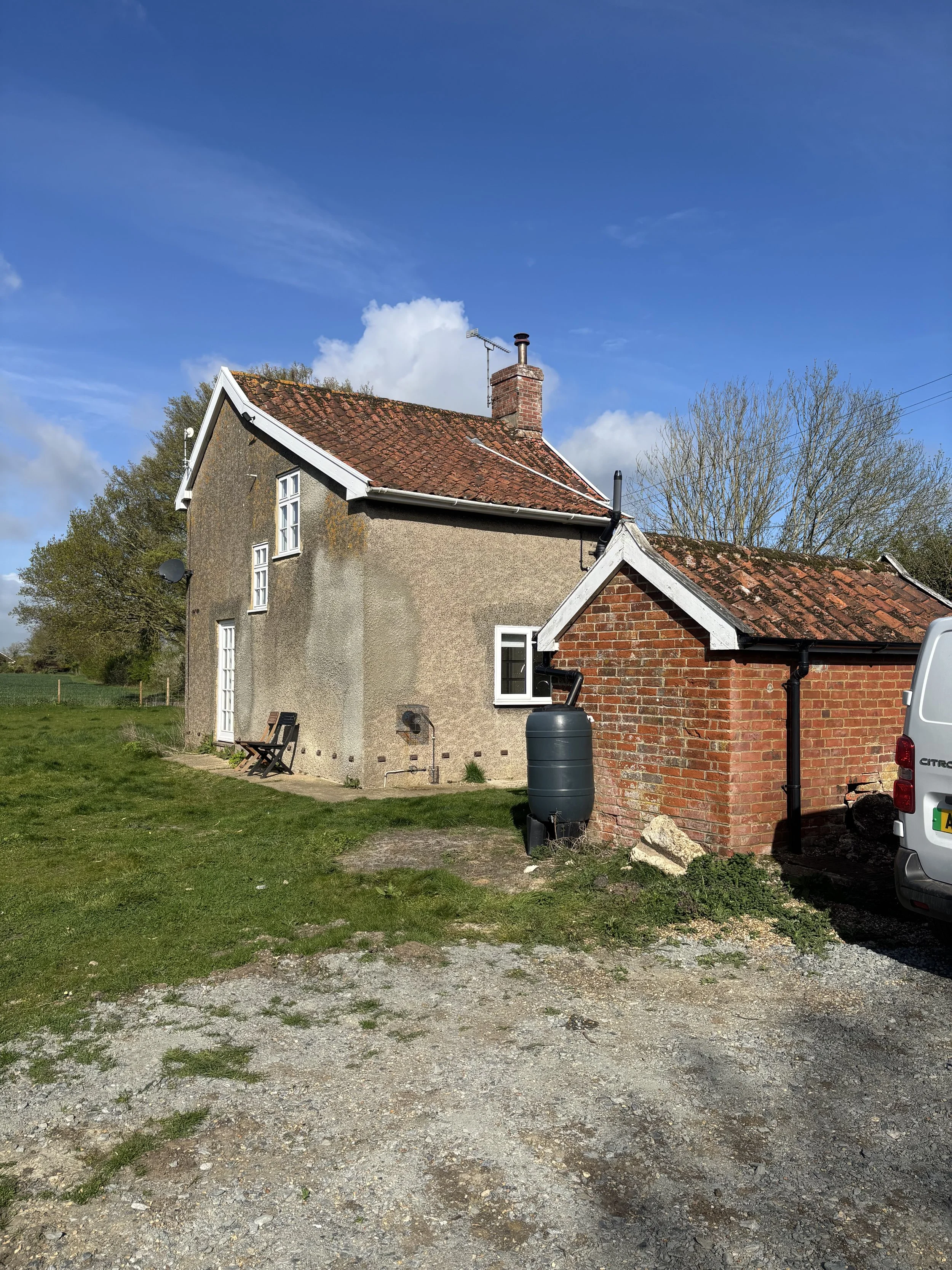 A house with a red brick chimney and a mix of stucco and brick exterior walls under a blue sky with some clouds. There is a grassy area with a gravel path in front, and a black rainwater barrel near the brick section of the house. A white van is part