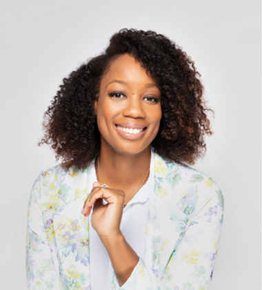 Portrait of a smiling woman with curly hair wearing a floral blazer over a white shirt against a light gray background.