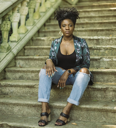 A woman sitting on outdoor stone steps, wearing a dark shirt, ripped jeans, a patterned jacket, and sandals.