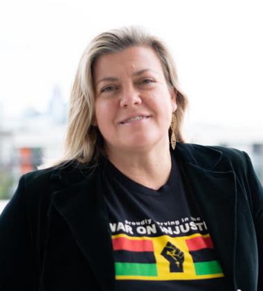 A woman with blonde hair wearing a black jacket and a black T-shirt with a colorful graphic, standing outdoors with a cityscape in the background.