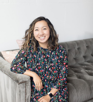 A woman smiling while sitting on a gray couch in a room with white walls, wearing a colorful floral dress and a smartwatch.