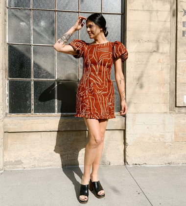 Woman in a short rust-colored dress with abstract white patterns, standing against a concrete wall with a large window.