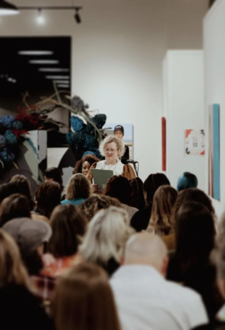 A woman with curly blonde hair holding a paper and standing at a microphone in front of a crowd of people in an art gallery or exhibition space.